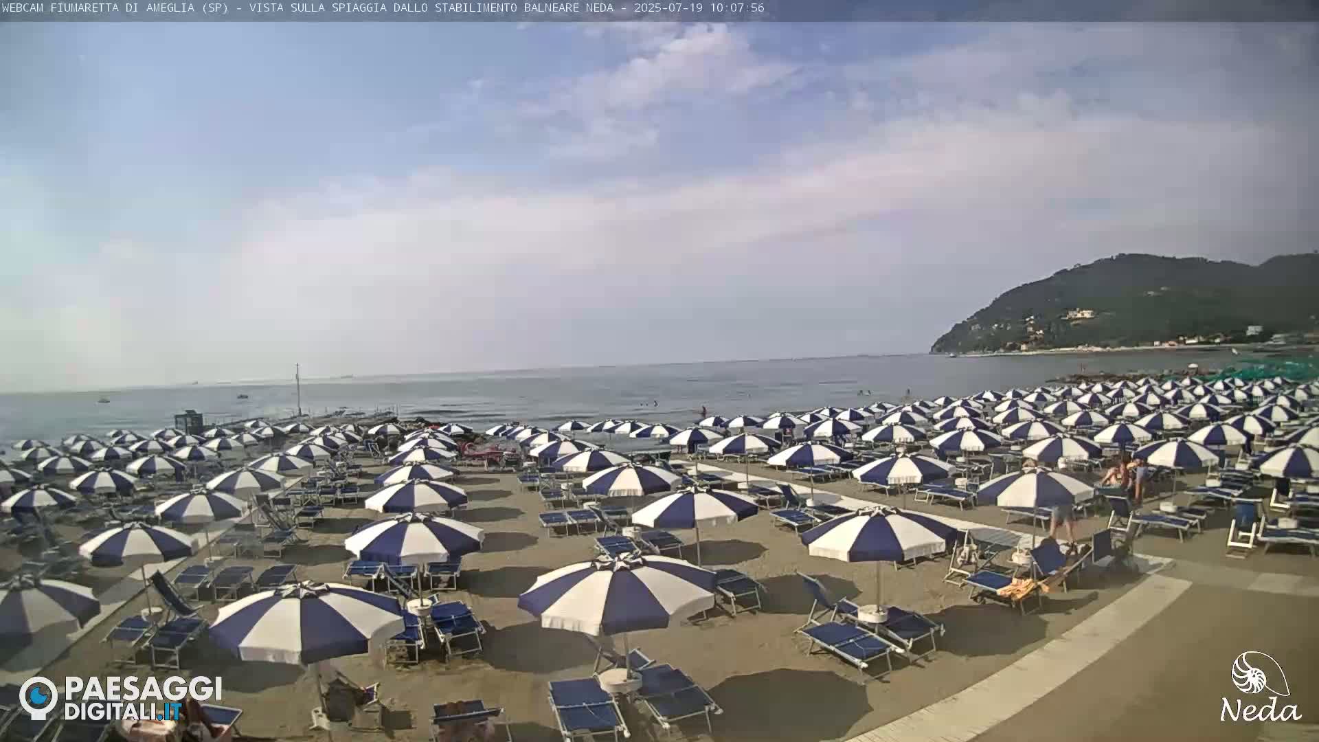 A sandy beach is densely populated with blue and white striped umbrellas and lounge chairs under a partly cloudy sky.