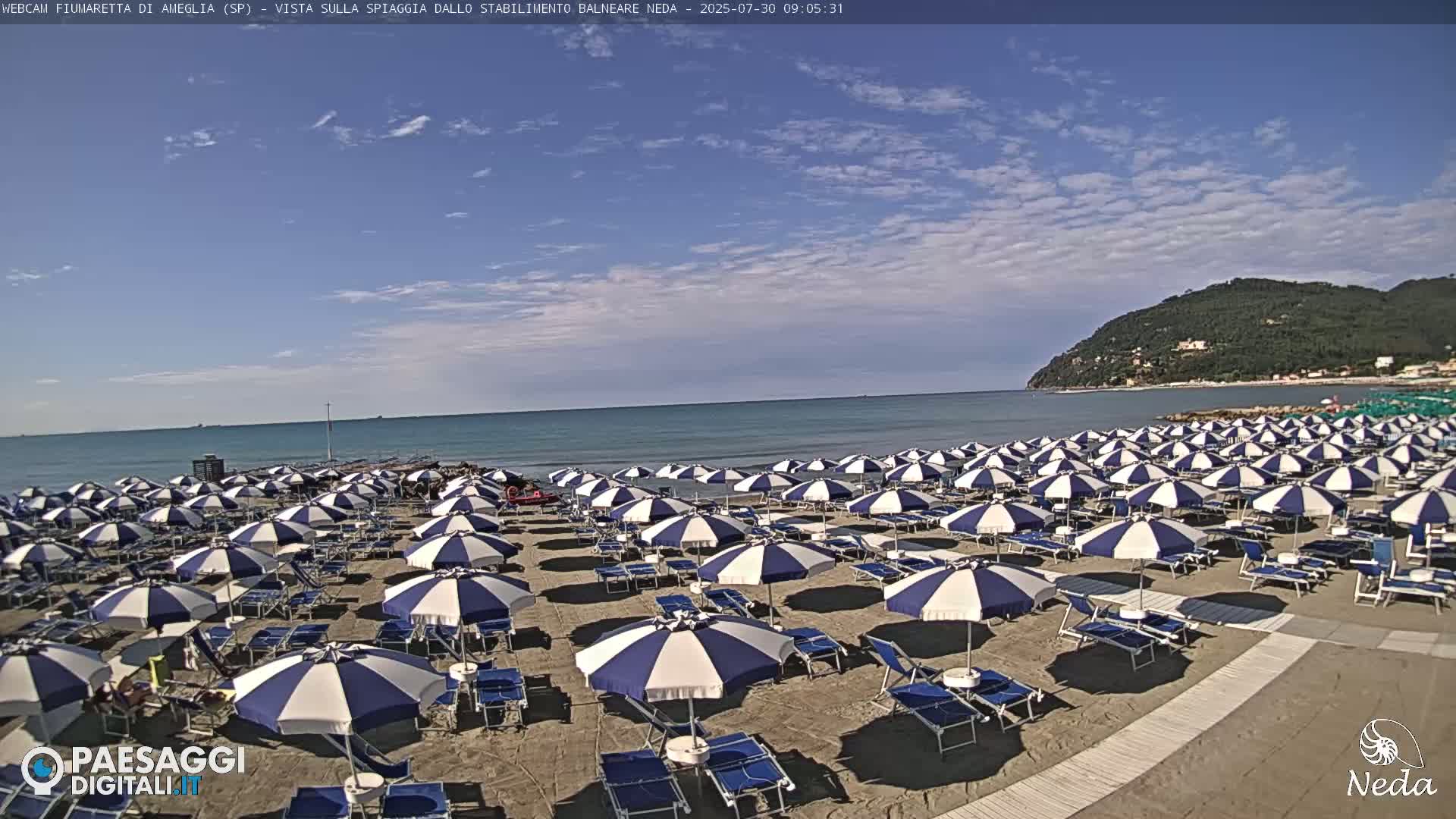 A sandy beach with numerous blue and white striped umbrellas under a mostly sunny sky.