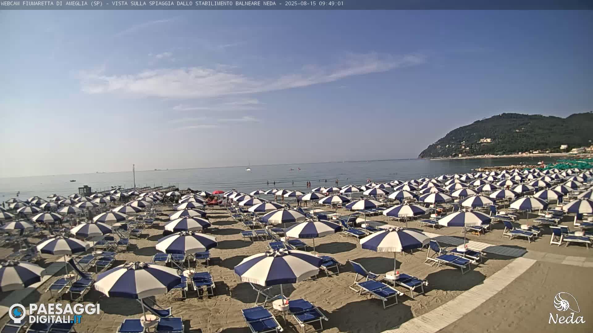 A sandy beach with numerous blue and white striped umbrellas and lounge chairs is shown under a mostly sunny sky, overlooking a calm sea with a distant hill in the background.