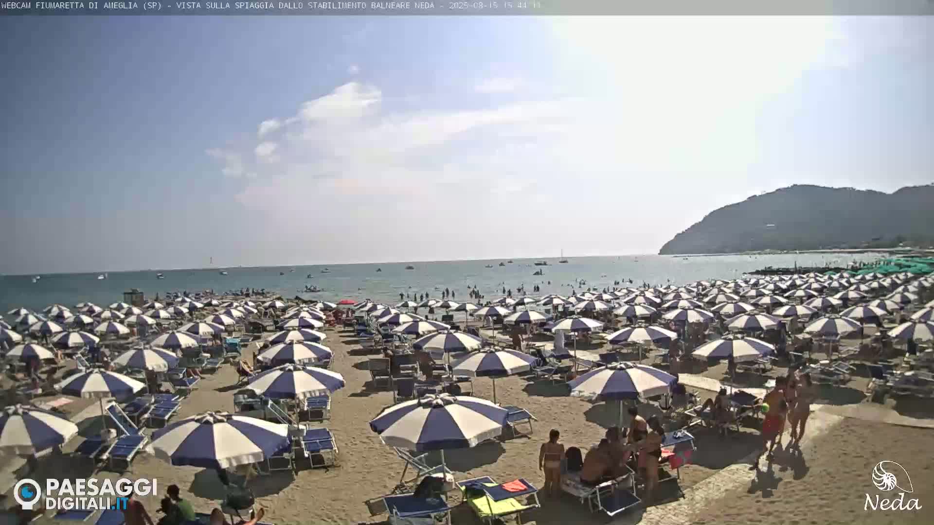 A sandy beach with numerous blue and white striped umbrellas and lounge chairs is shown under a mostly sunny sky, overlooking a calm sea with a distant hill in the background.