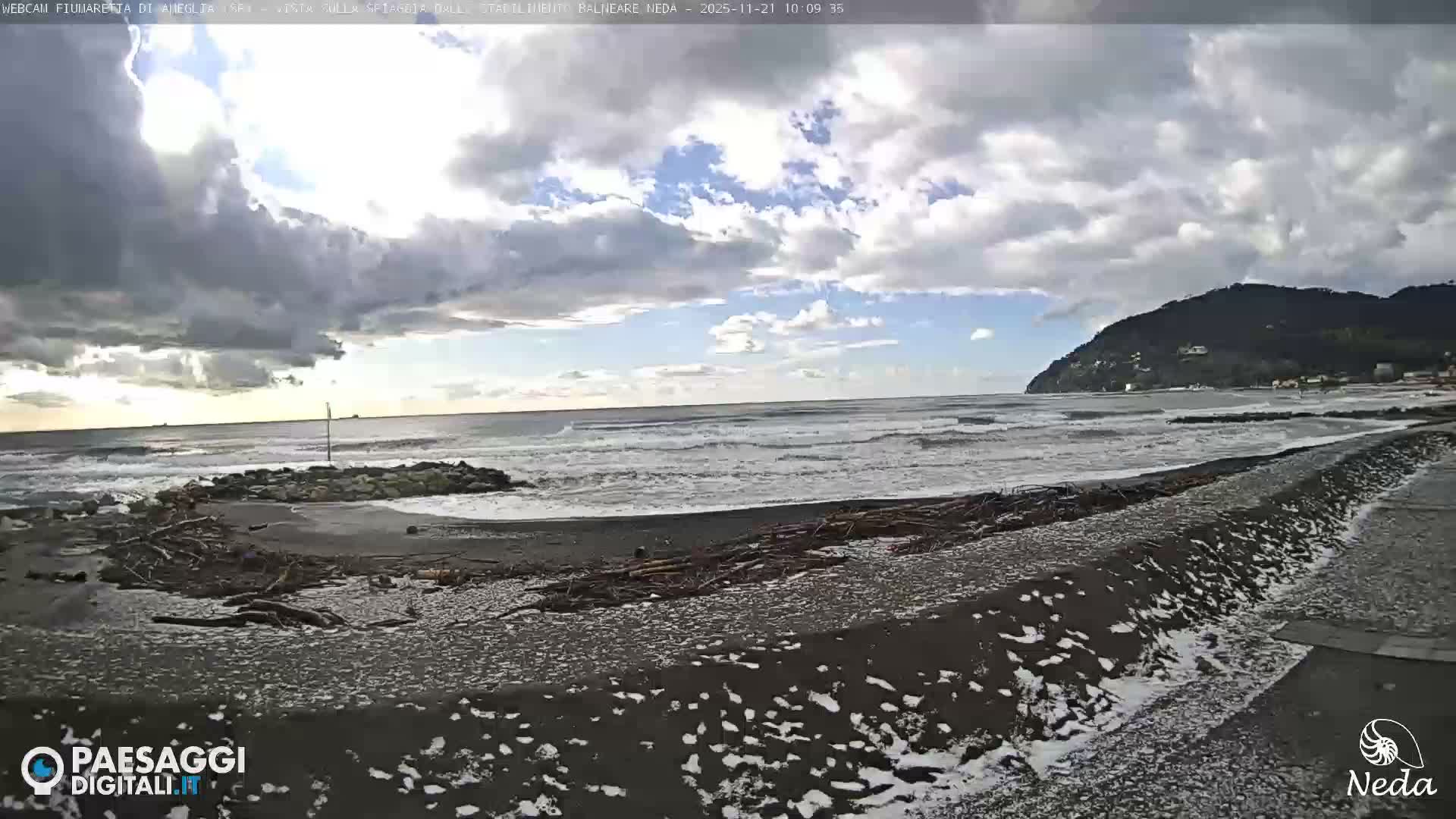 The image shows a partly cloudy day at a dark-pebbled beach with moderate waves breaking, extensive driftwood scattered along the shore, a rocky jetty, and a green, hilly coastline dotted with buildings in the background.