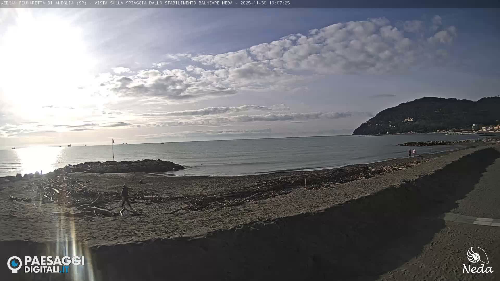A bright, partly cloudy day illuminates a wide beach scattered with driftwood, where a person walks along the shore facing a calm sea with distant boats, flanked on the right by a forested hillside.
