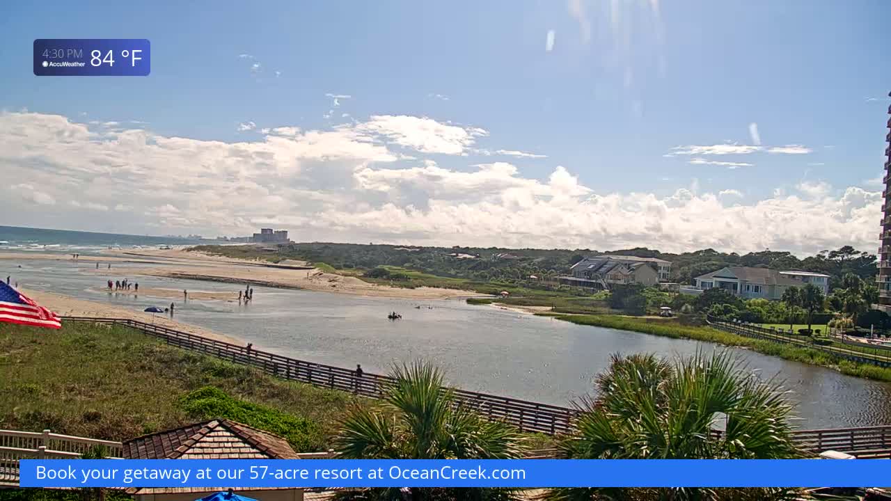 A partly cloudy, 84°F day shows an inlet flowing from a beach to a calm lagoon, lined with a wooden boardwalk and bordered by homes and vegetation.