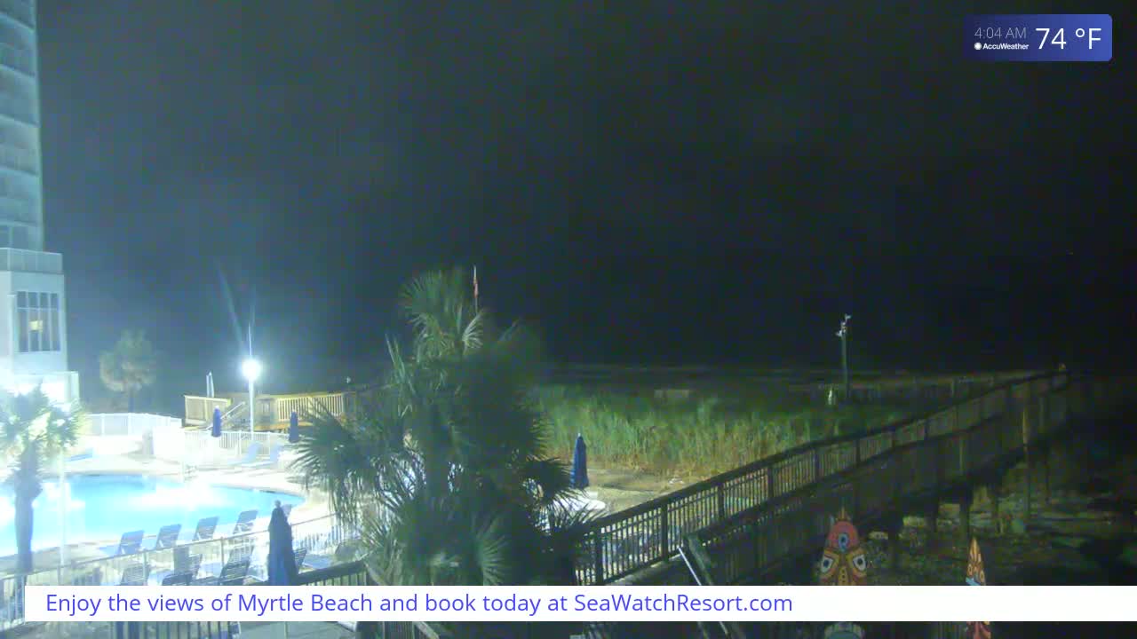 A nighttime view of a resort pool area, palm trees, and a boardwalk leading to a dark beach under a clear, 74°F sky.