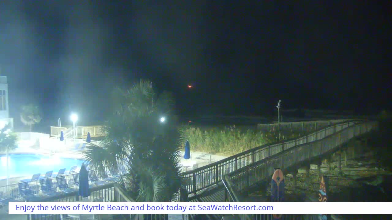 On a clear night, a resort scene features a brightly lit swimming pool and palm trees on the left, leading to a wooden boardwalk that stretches over grassy dunes towards a dark ocean horizon, with a faint reddish crescent visible in the vast night sky.
