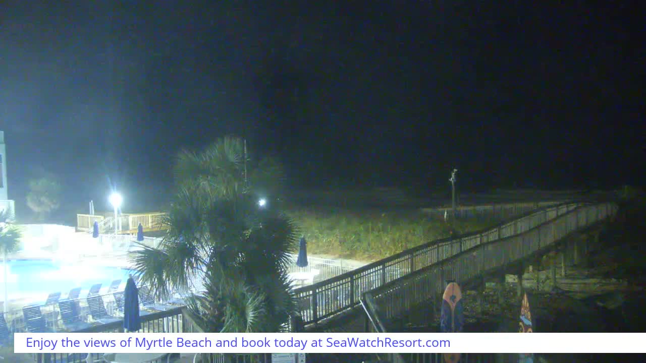 An illuminated resort swimming pool with lounge chairs and palm trees is visible at night, alongside a wooden boardwalk leading towards a dark, grassy beach or dune area under a clear sky.