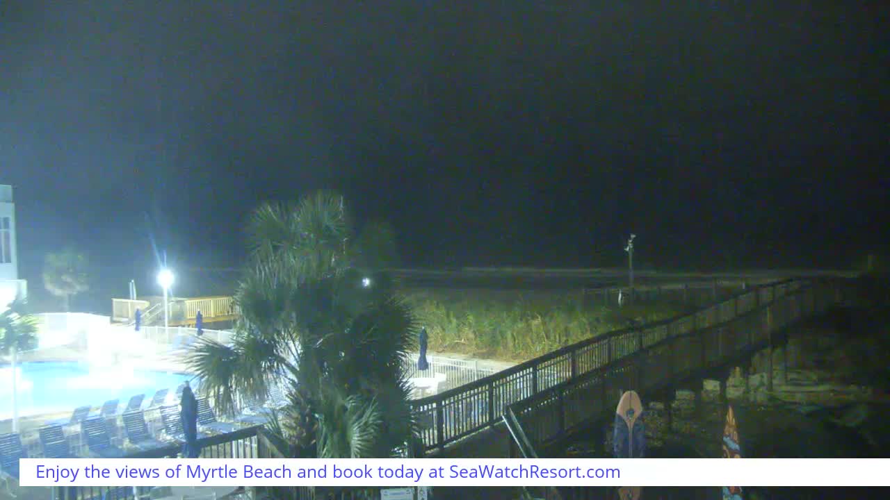 A dark, overcast night view from a beachfront resort reveals an illuminated swimming pool with lounge chairs, palm trees, and a wooden boardwalk extending towards a dimly visible ocean.