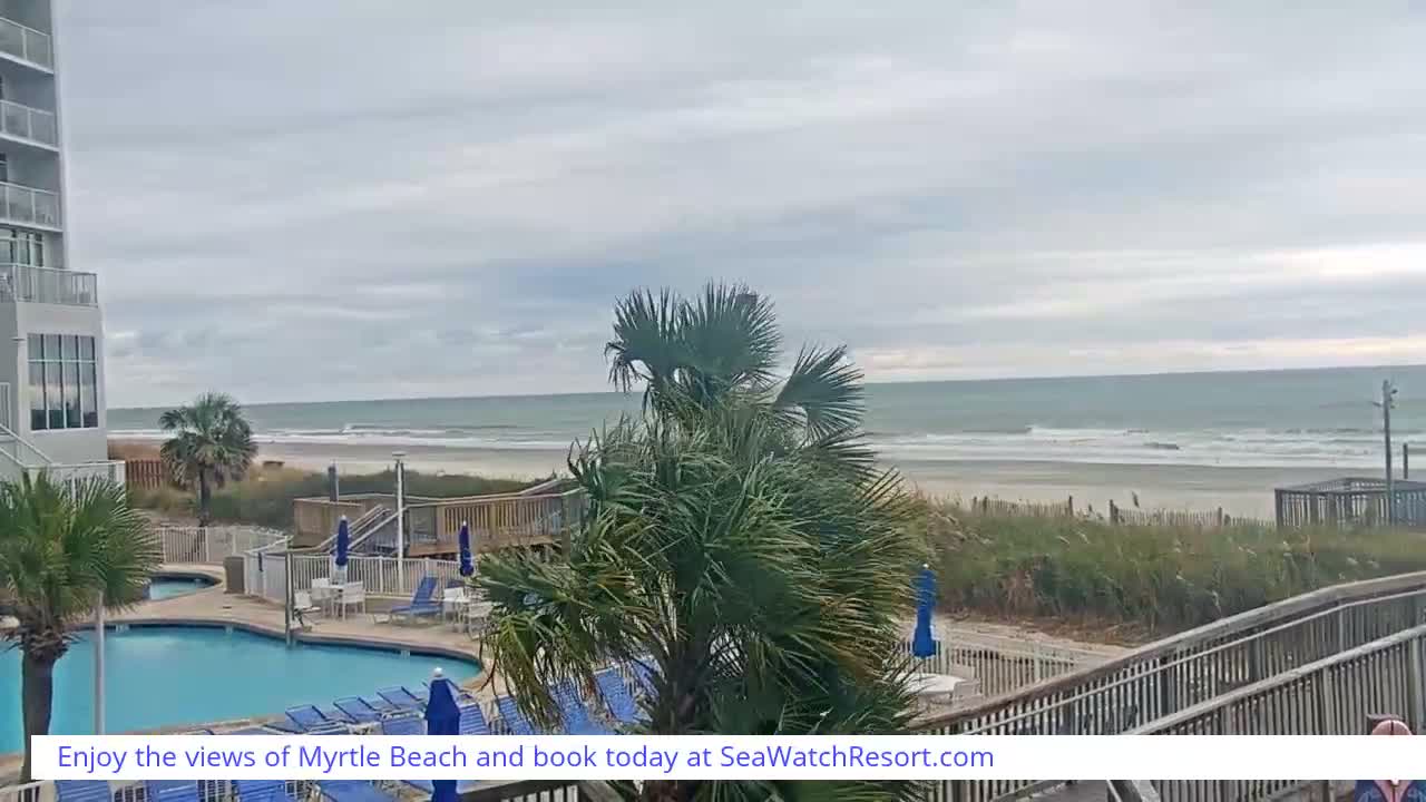 A dark, overcast night view from a beachfront resort reveals an illuminated swimming pool with lounge chairs, palm trees, and a wooden boardwalk extending towards a dimly visible ocean.