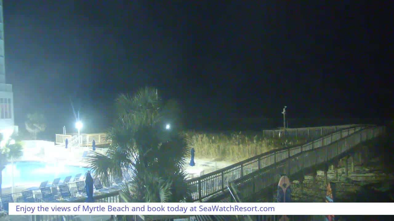 A dark, overcast night view from a beachfront resort reveals an illuminated swimming pool with lounge chairs, palm trees, and a wooden boardwalk extending towards a dimly visible ocean.
