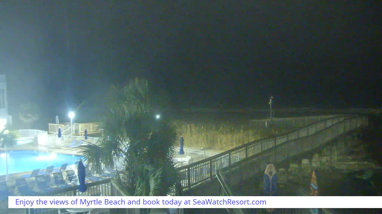A resort's illuminated swimming pool, palm trees, and a long boardwalk stretching towards the dark ocean are visible on a wet and hazy night.