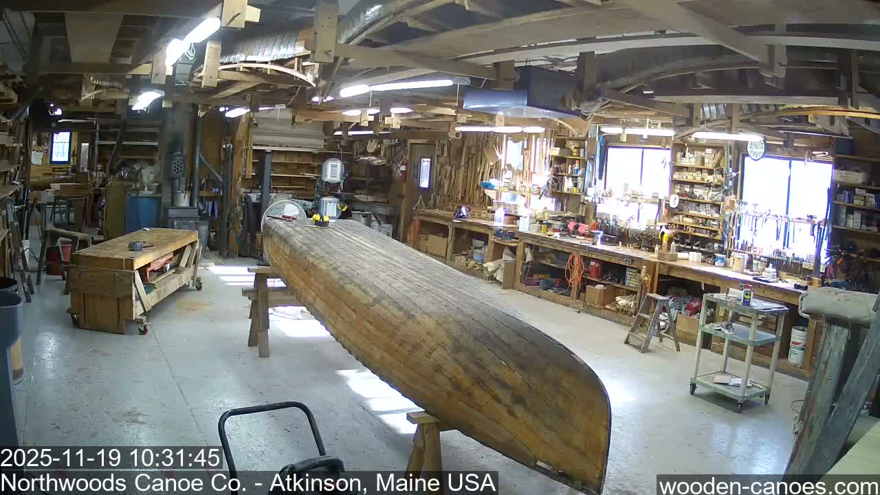 A beautifully crafted wooden canoe with a dark blue exterior and natural wood interior, complete with woven seats, rests on a stand in a sunny, grassy area with trees in the background.