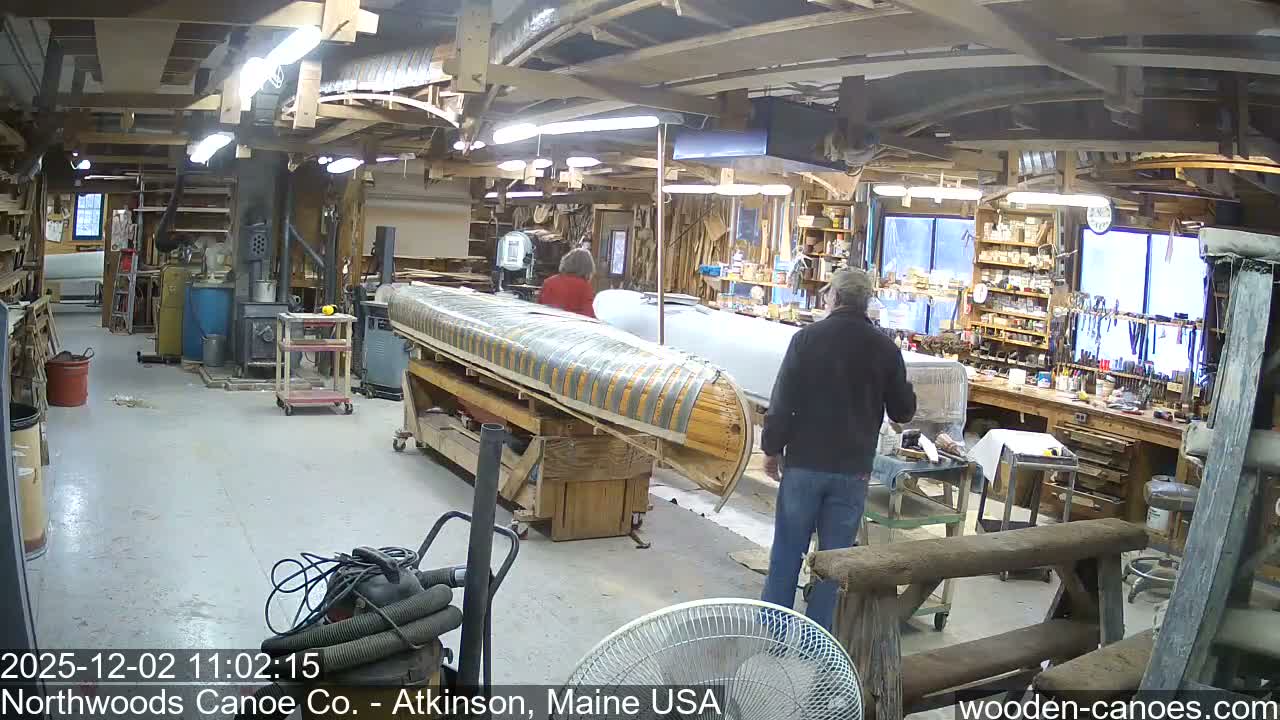 A brightly lit, bustling indoor woodworking shop is shown, featuring two individuals near a partially constructed wooden canoe hull on a stand, surrounded by tools and equipment, under bright daylight conditions.