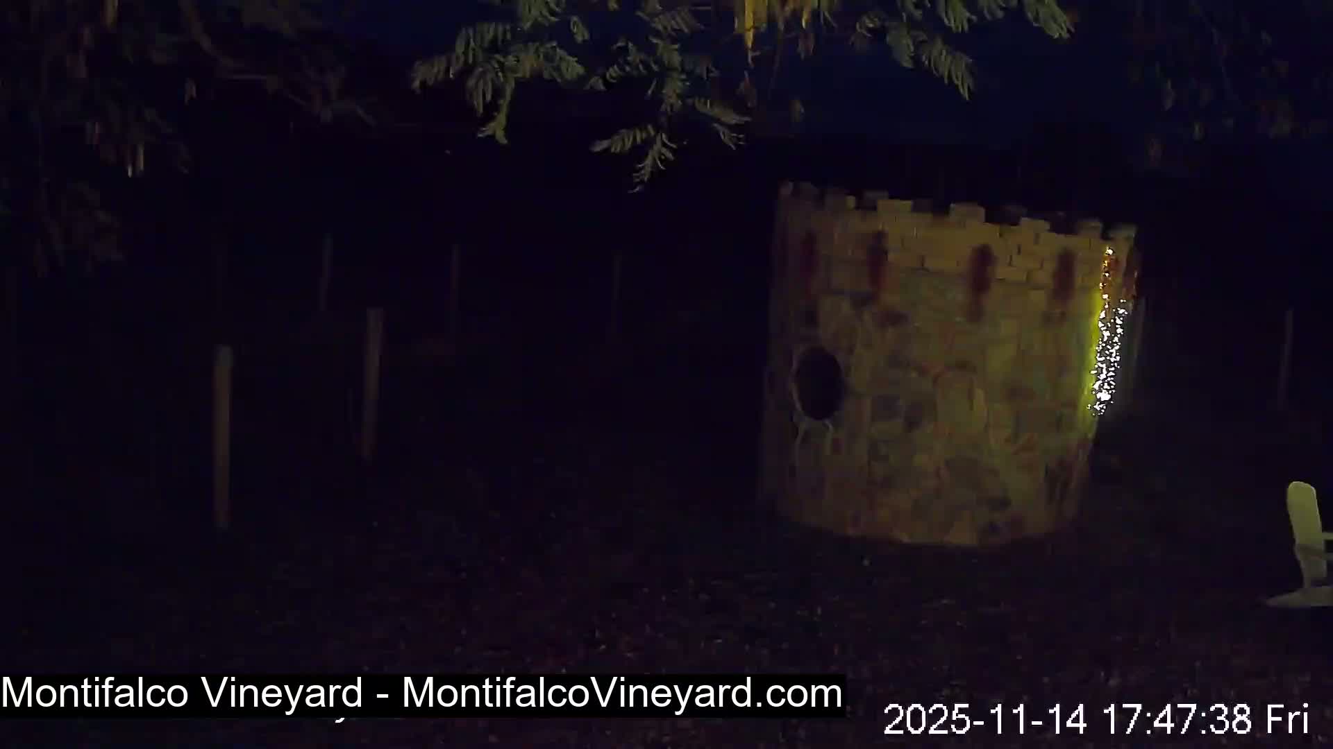 At night, a dimly lit, crenellated stone tower-like structure adorned with a string of lights and featuring a circular opening stands in an outdoor area alongside faint vineyard posts, dark foliage, and a partially visible chair under clear skies.