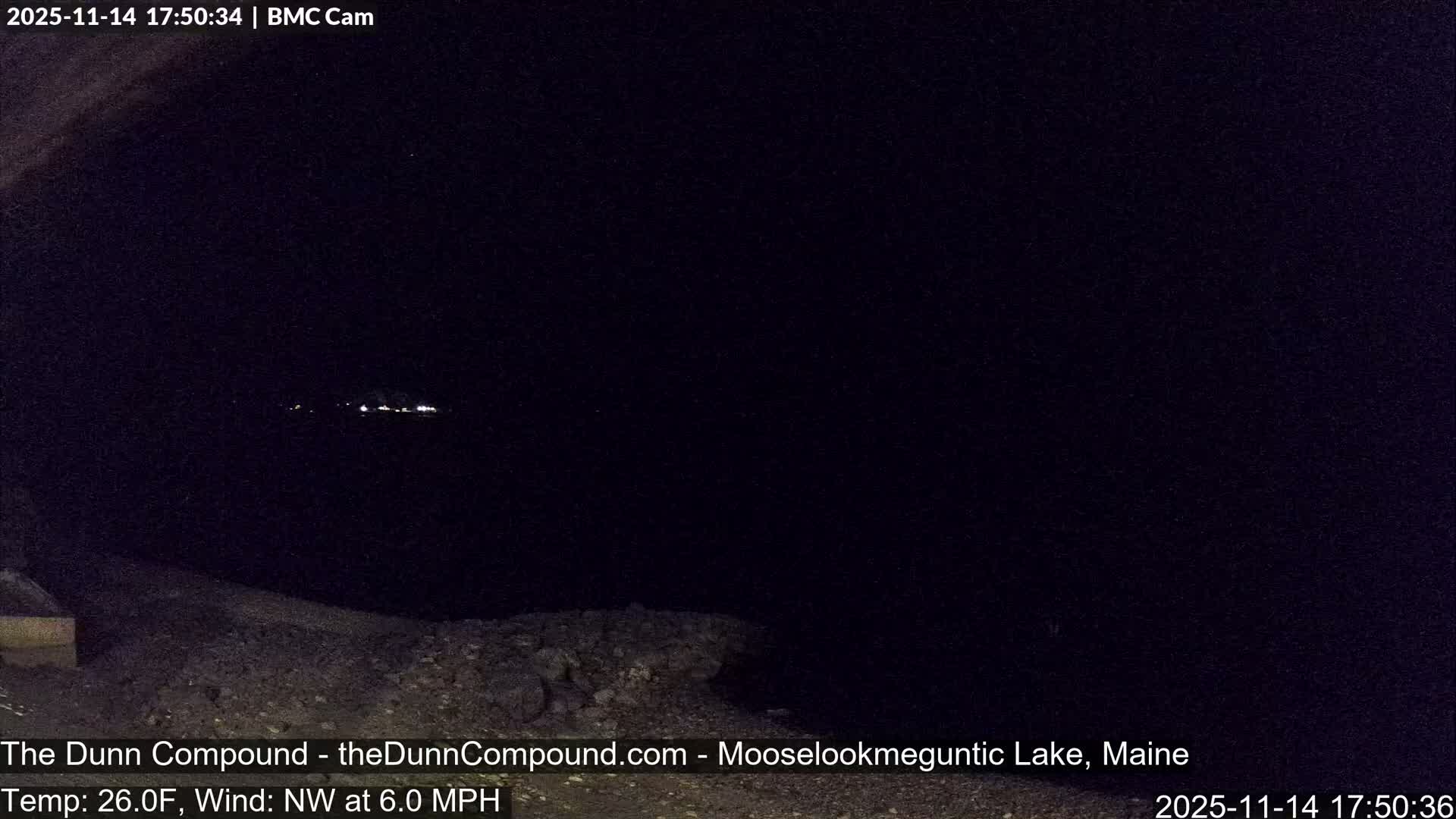 A very dark outdoor nighttime scene displays faint distant lights across a body of water with an indistinct foreground, experiencing a temperature of 26.0F and a 6.0 MPH northwest wind.