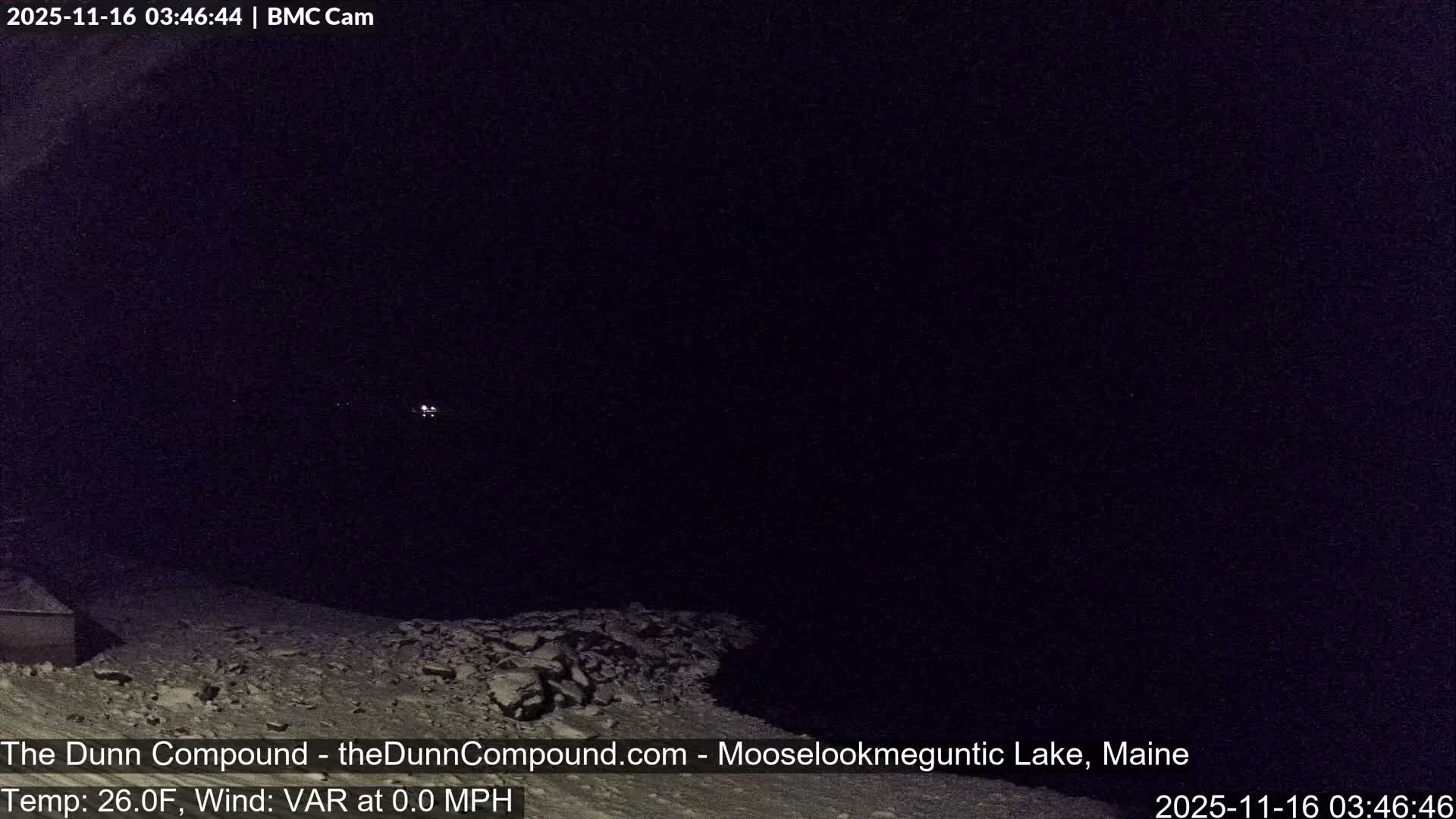 A dark nighttime scene reveals a snow-covered or rocky shore with a small structure to the left, bordering a vast dark expanse that holds a few faint lights in the distance, all under clear, calm, and cold conditions.