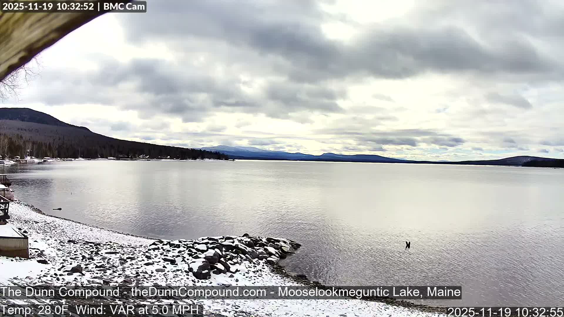 A dark nighttime scene reveals a snow-covered or rocky shore with a small structure to the left, bordering a vast dark expanse that holds a few faint lights in the distance, all under clear, calm, and cold conditions.