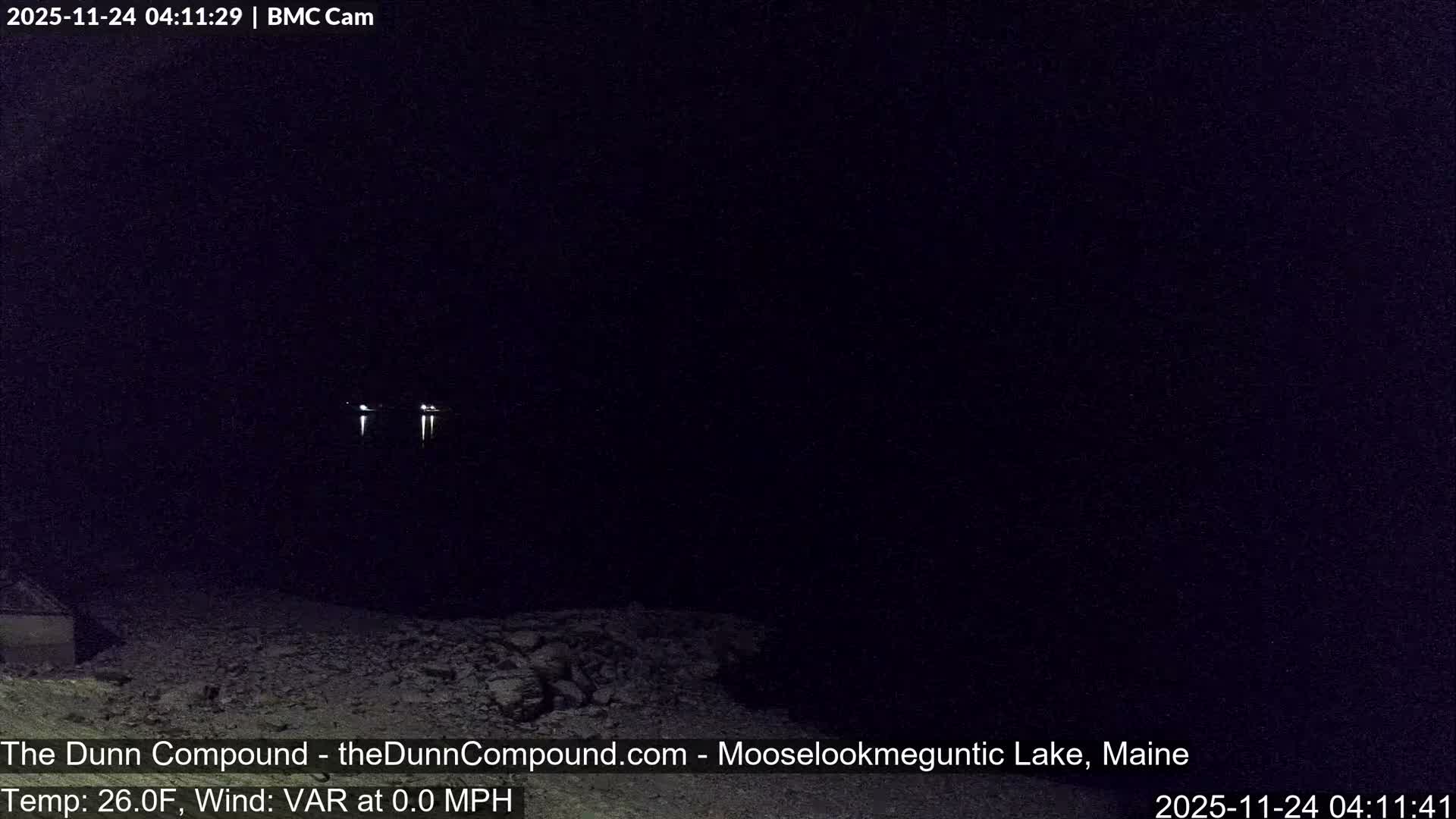 A dark nighttime scene reveals faint lights reflecting on a calm body of water, with a rocky foreground, under clear conditions.