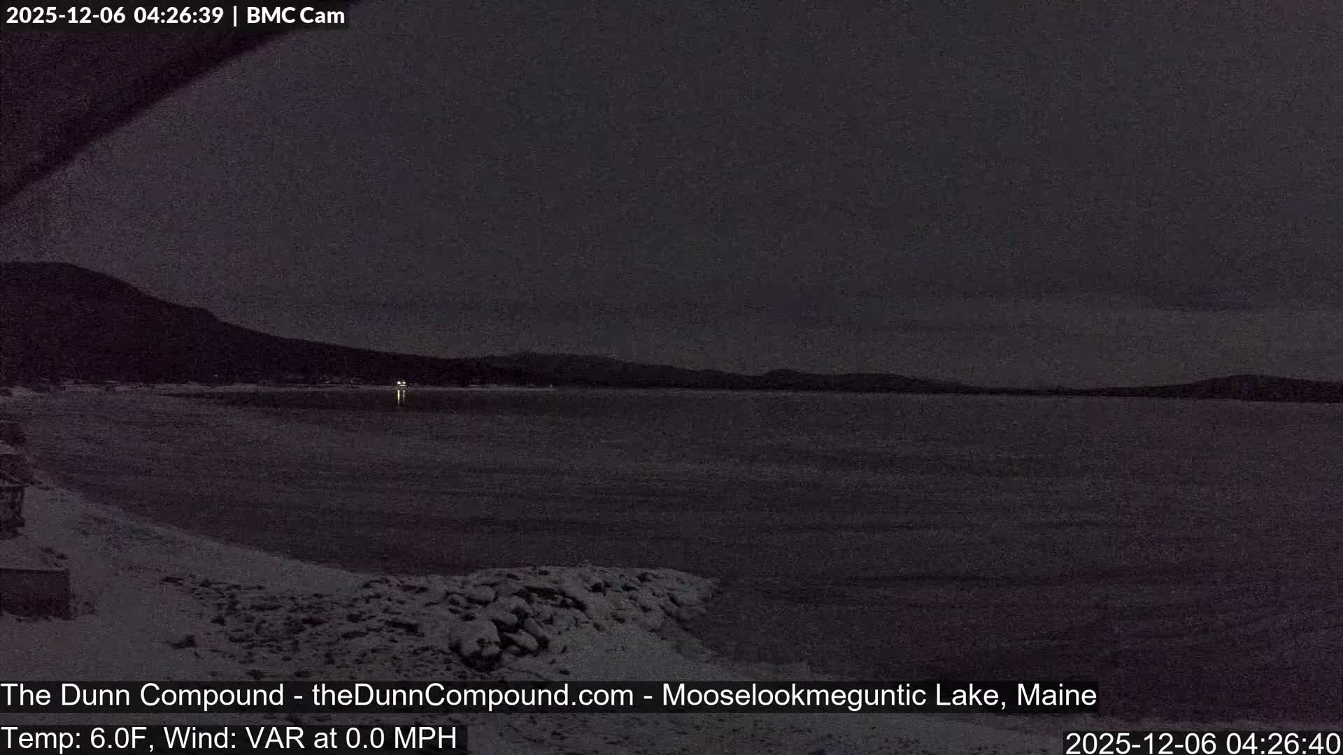 A dark, low-light winter scene reveals a calm lake reflecting a few distant lights, with a snowy or icy shoreline and dark hills under a heavily overcast night sky.