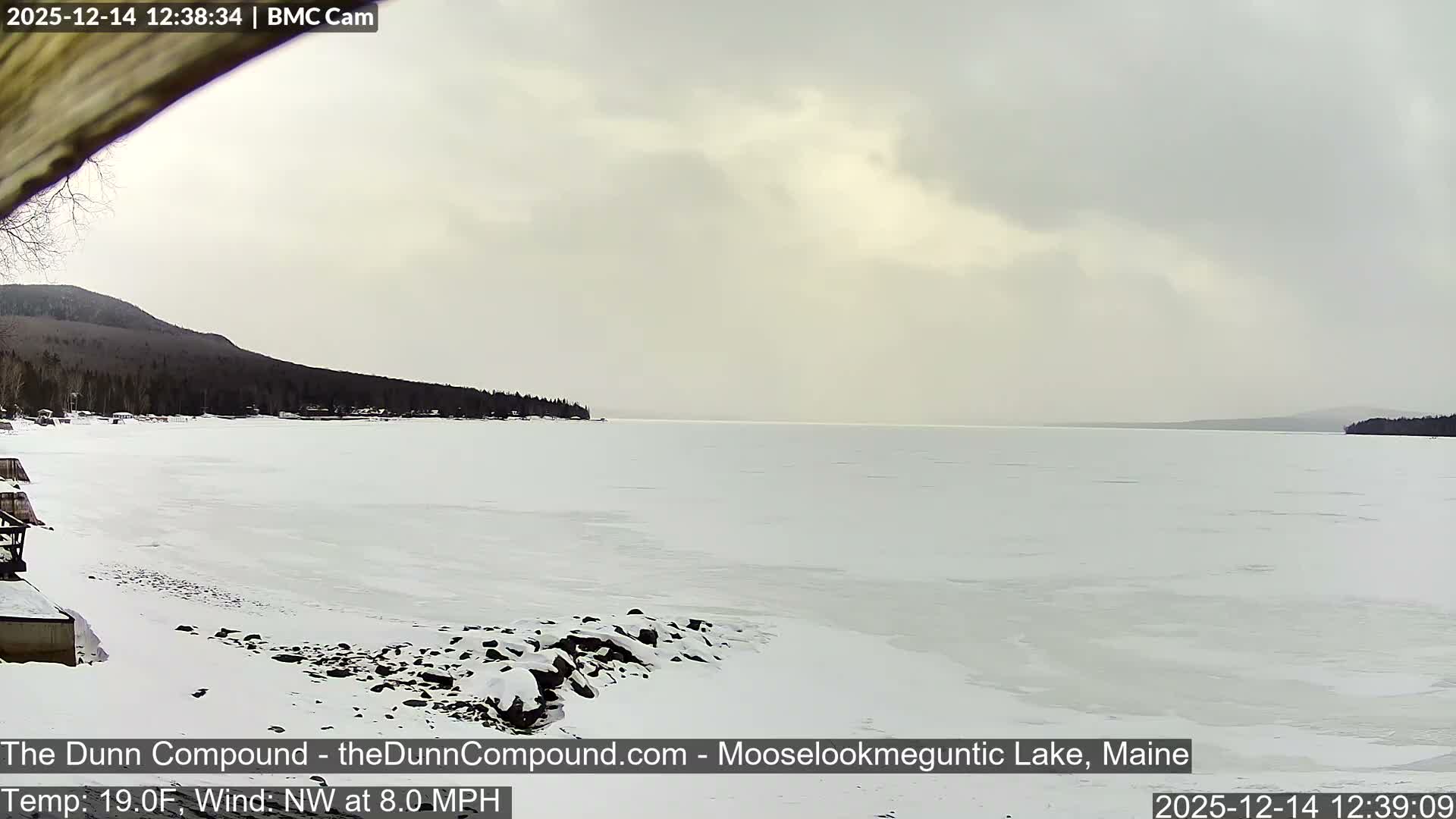 A dark, low-light winter scene reveals a calm lake reflecting a few distant lights, with a snowy or icy shoreline and dark hills under a heavily overcast night sky.
