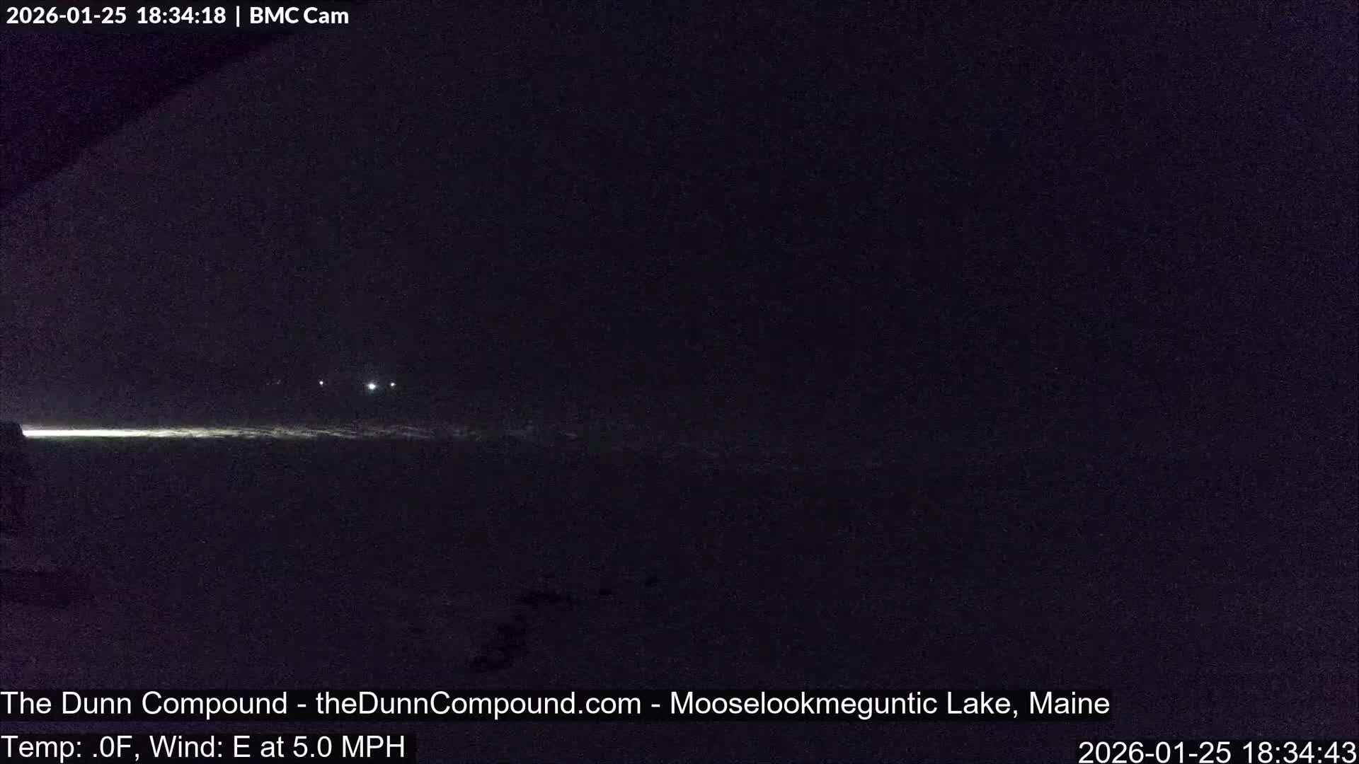 A dark outdoor scene at night reveals a bright horizontal light on the lower left illuminating a likely snow-covered or icy foreground, with several faint lights visible in the distant background under a clear, dark sky.