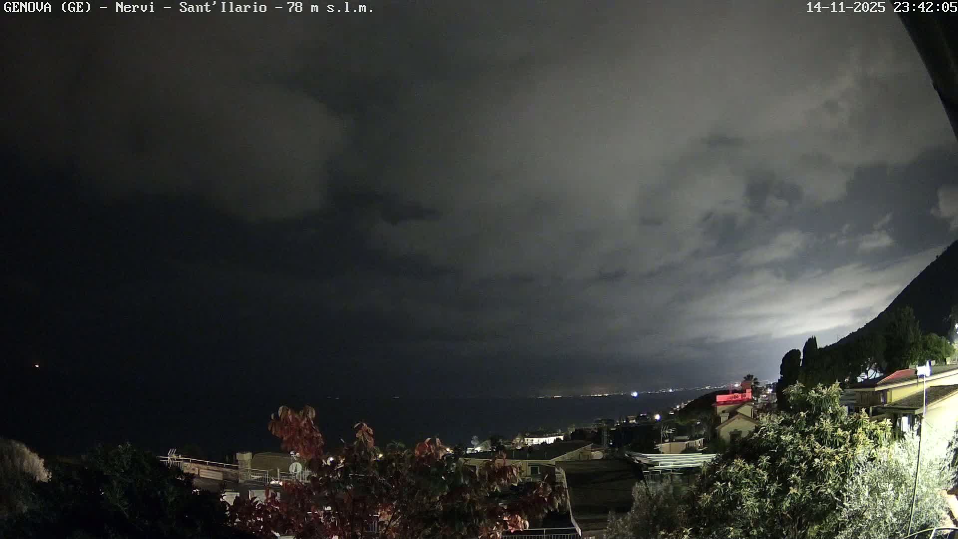 The image shows a dark, cloudy night over a coastal town with scattered lights illuminating buildings and the distant shoreline, with a dark mountain rising on the right.