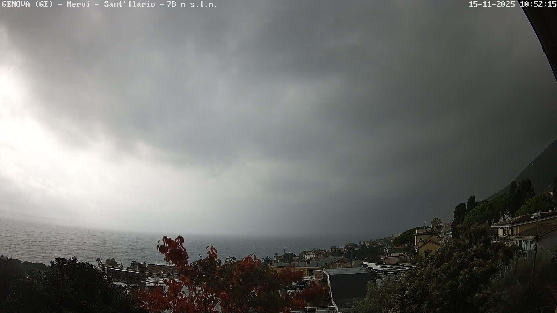 A dark, heavily overcast sky looms over a choppy sea and a coastal town, with autumn-colored trees and a steep, tree-covered hillside framing the scene on what appears to be a stormy day.