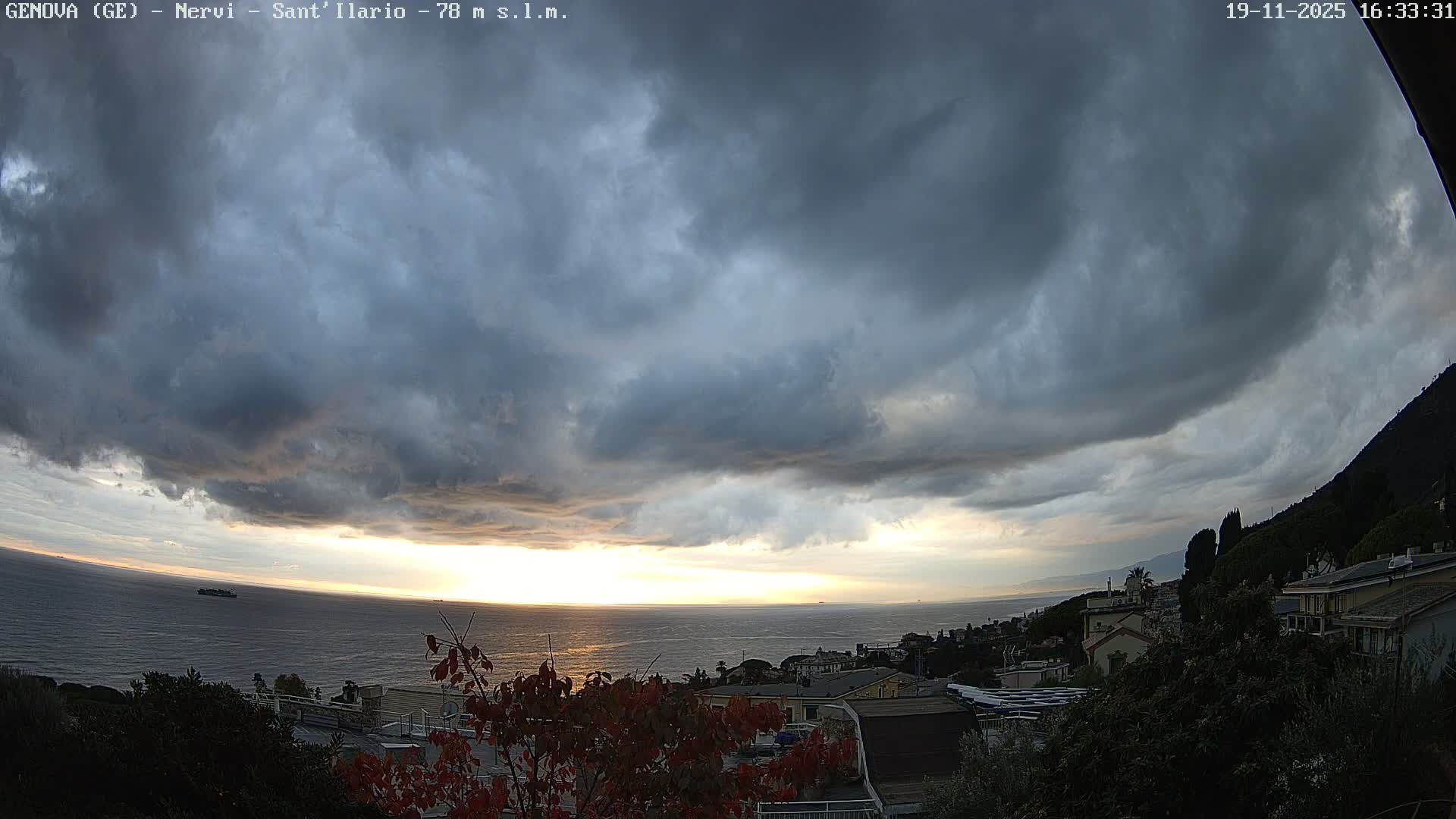 An overcast, grey sky hangs over a coastal town nestled between lush hills and a dark sea, with trees in the foreground displaying touches of autumn color.