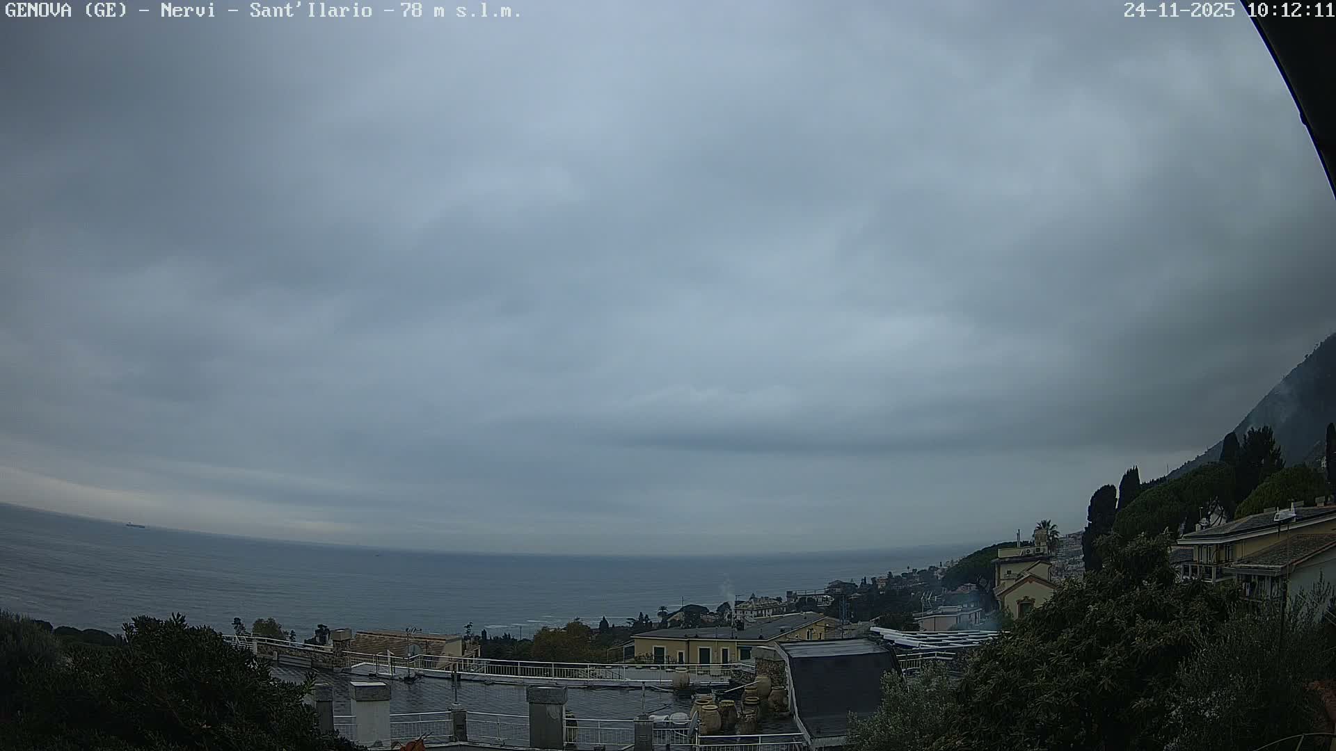 An overcast sky hangs over a coastal town featuring multi-level buildings, lush trees, and a steep, forested hillside, with the grey sea extending to the horizon where a distant ship sails.