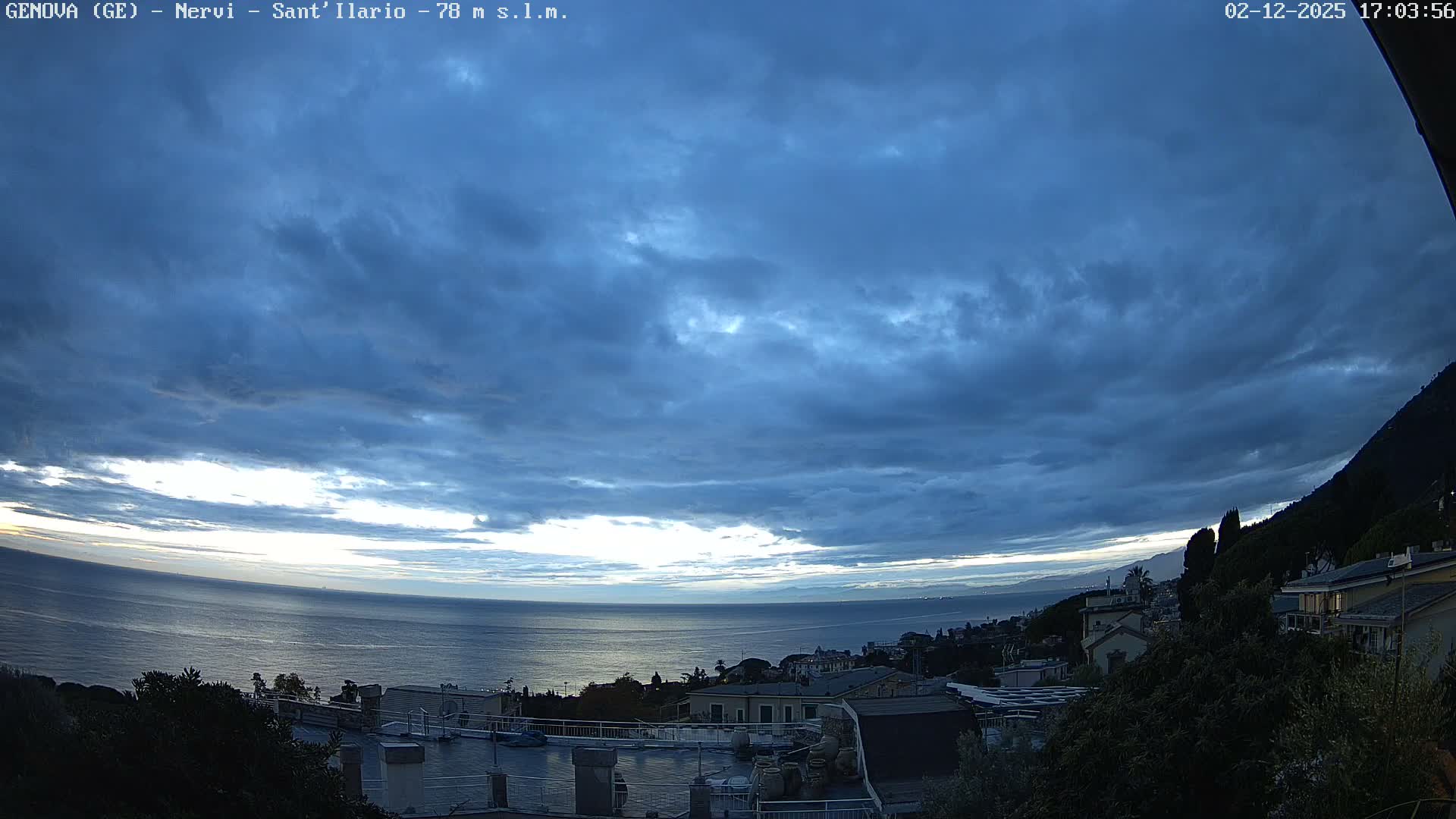 Under a heavily overcast, dark blue-grey sky with a bright sliver of light on the horizon, a coastal landscape features a calm sea, a town built along the shore with visible rooftops and lush vegetation, and a steep, tree-covered mountain rising on the right.