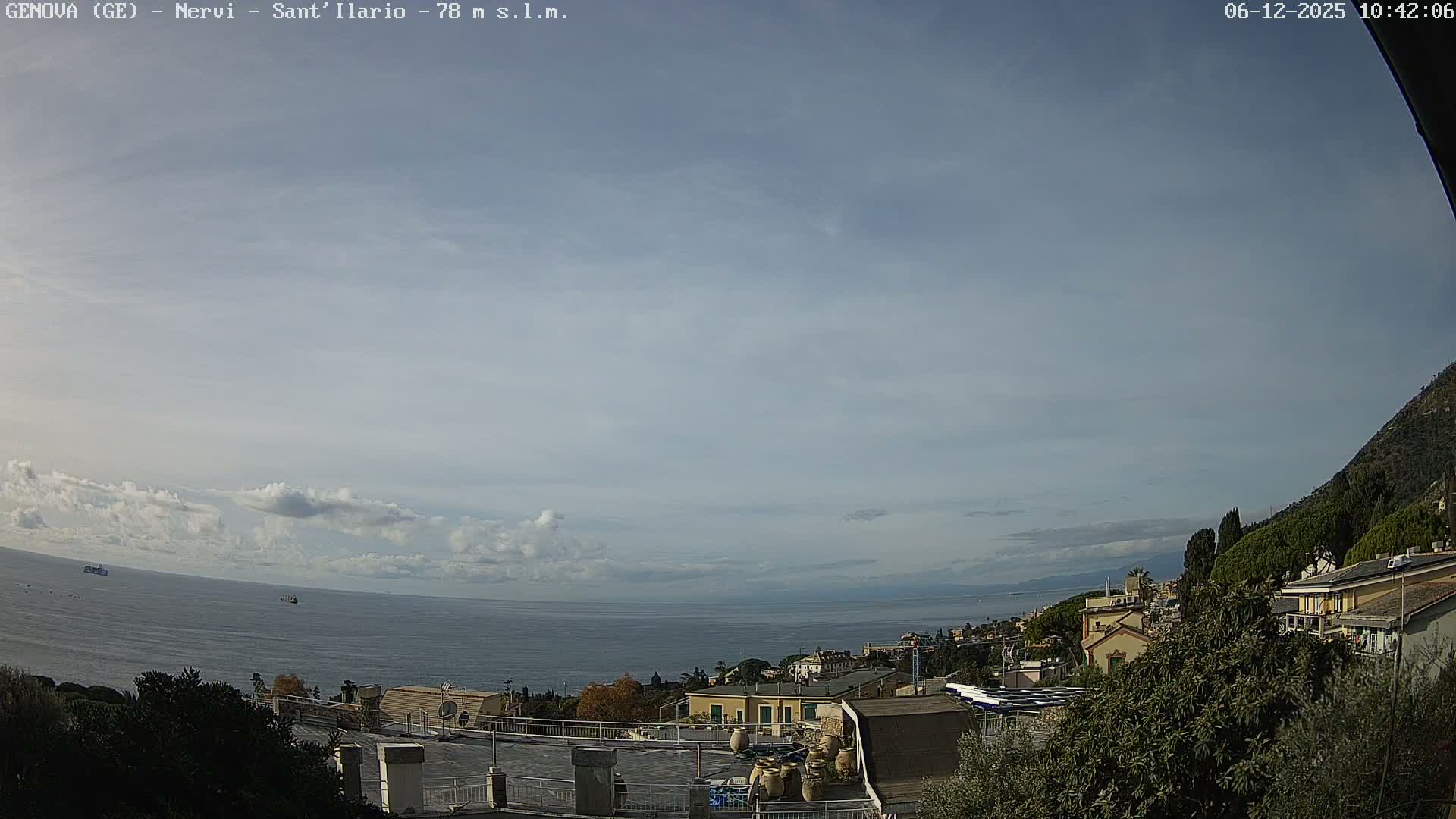 A wide view captures a coastal town with buildings nestled on a green hillside overlooking a calm sea dotted with two ships, all under a partly cloudy sky.
