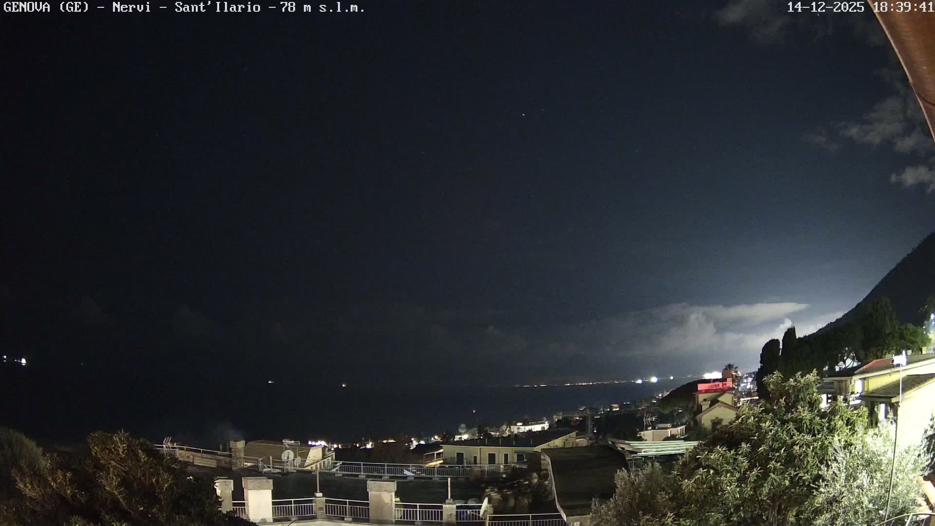 A wide view captures a coastal town with buildings nestled on a green hillside overlooking a calm sea dotted with two ships, all under a partly cloudy sky.