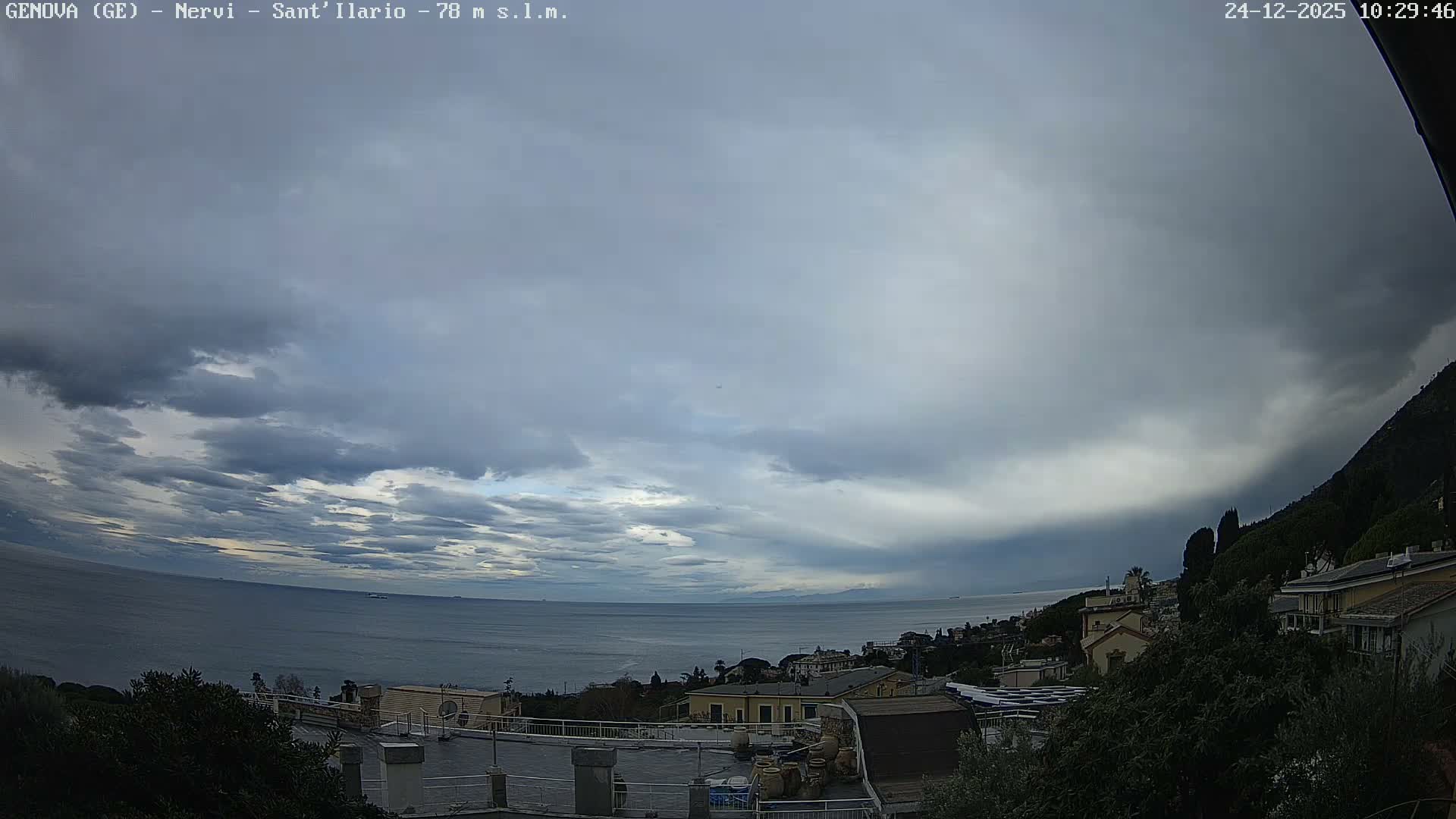 A wide view captures a coastal town with buildings nestled on a green hillside overlooking a calm sea dotted with two ships, all under a partly cloudy sky.