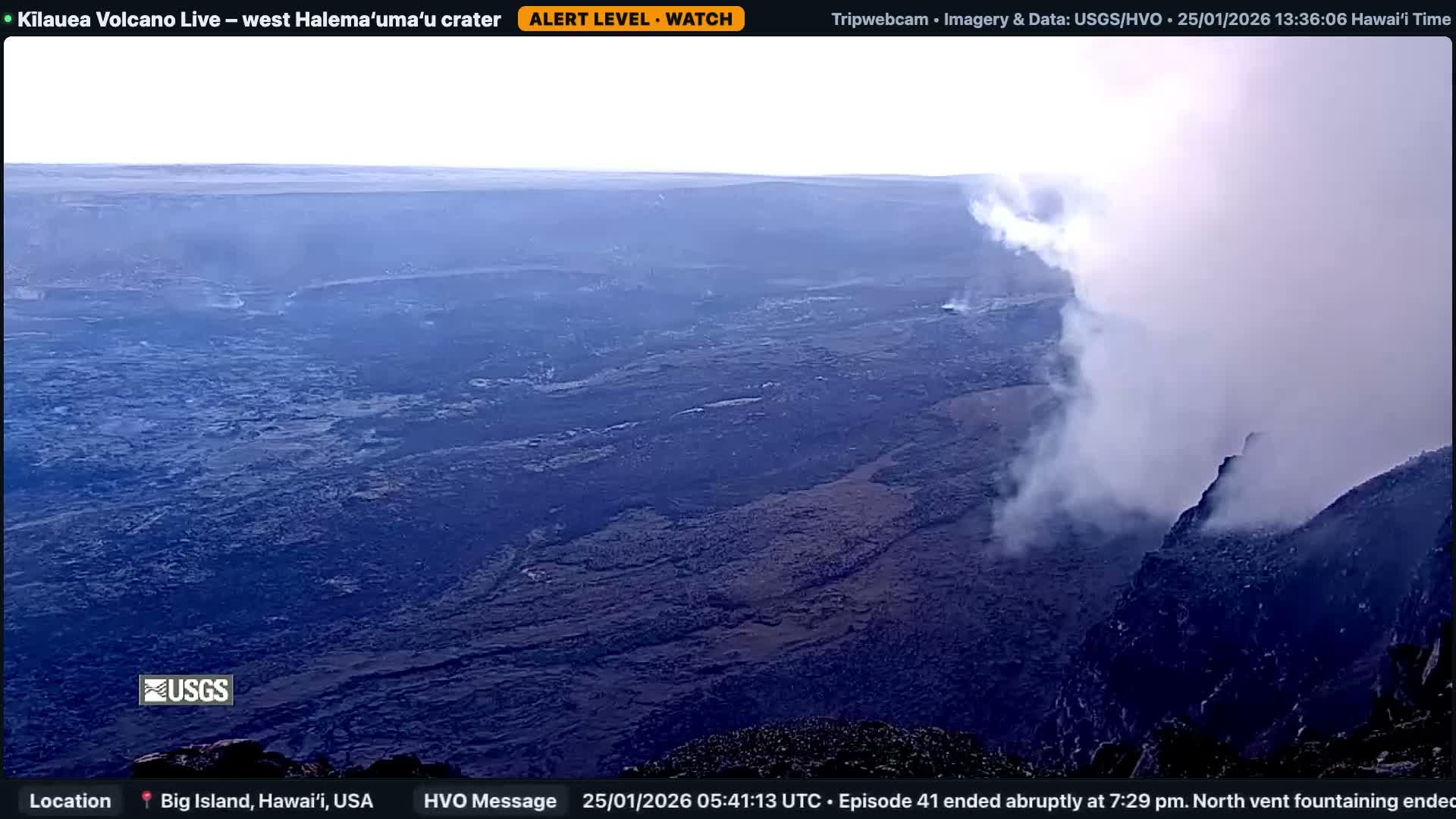 The image shows a vast, dark, volcanic caldera floor with a prominent plume of white steam or gas rising from a vent on the right side, all under a clear, bright sky.