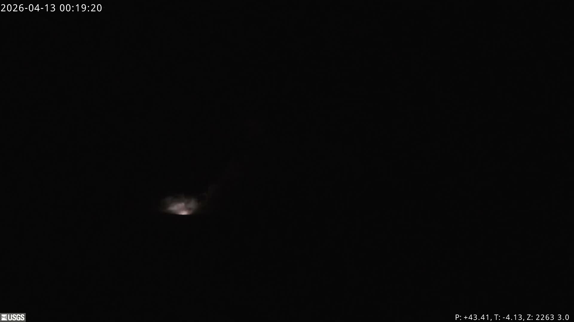 At night, a dark volcanic landscape is illuminated by brightly glowing areas on the left, with white and gray steam plumes rising and drifting across the terrain under clear sky conditions.