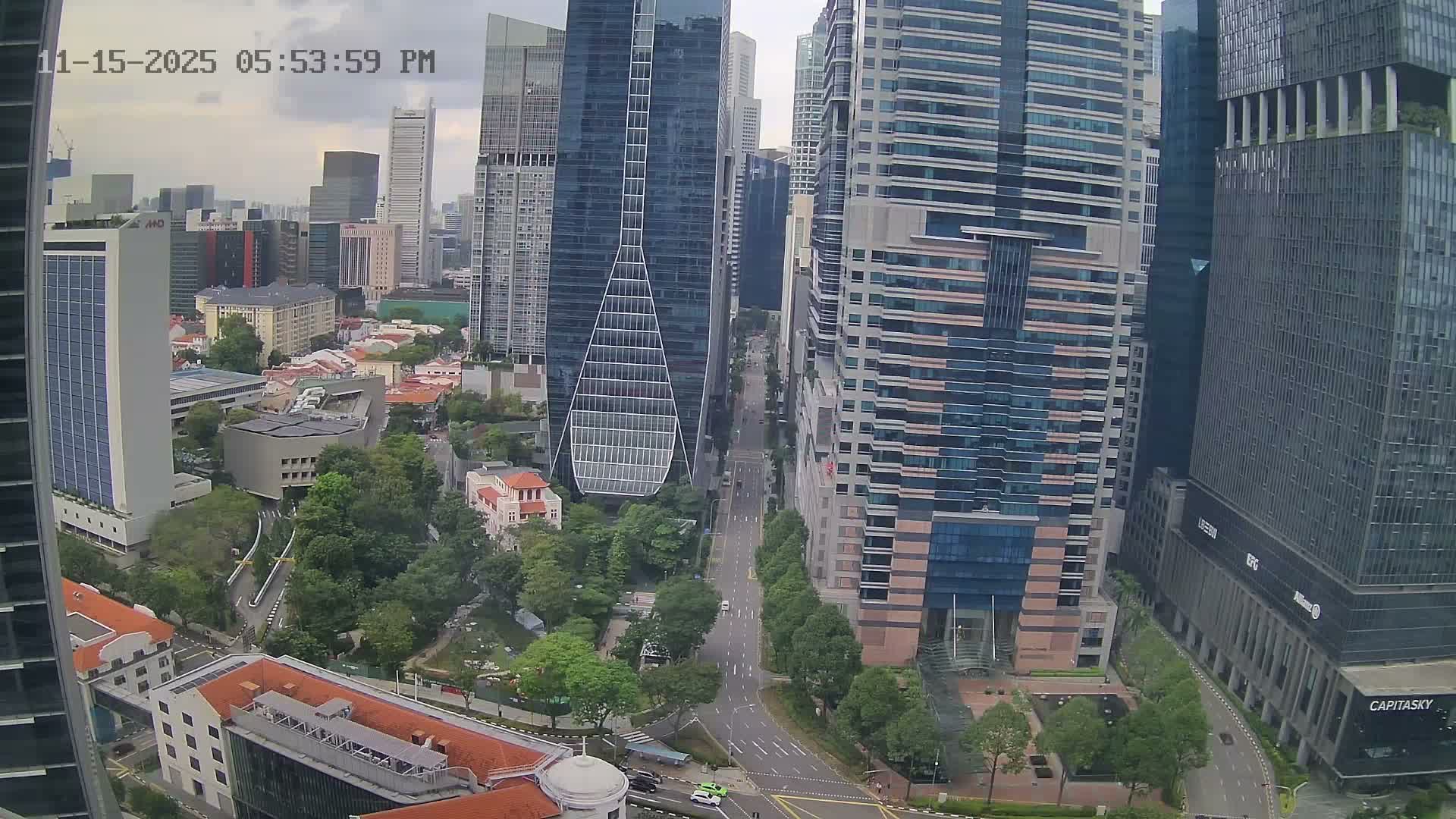 An elevated view captures a sprawling modern city featuring numerous skyscrapers, including a distinctive triangular-shaped glass building, alongside tree-lined streets, light vehicle traffic, and a few traditional low-rise buildings, all under an overcast sky.