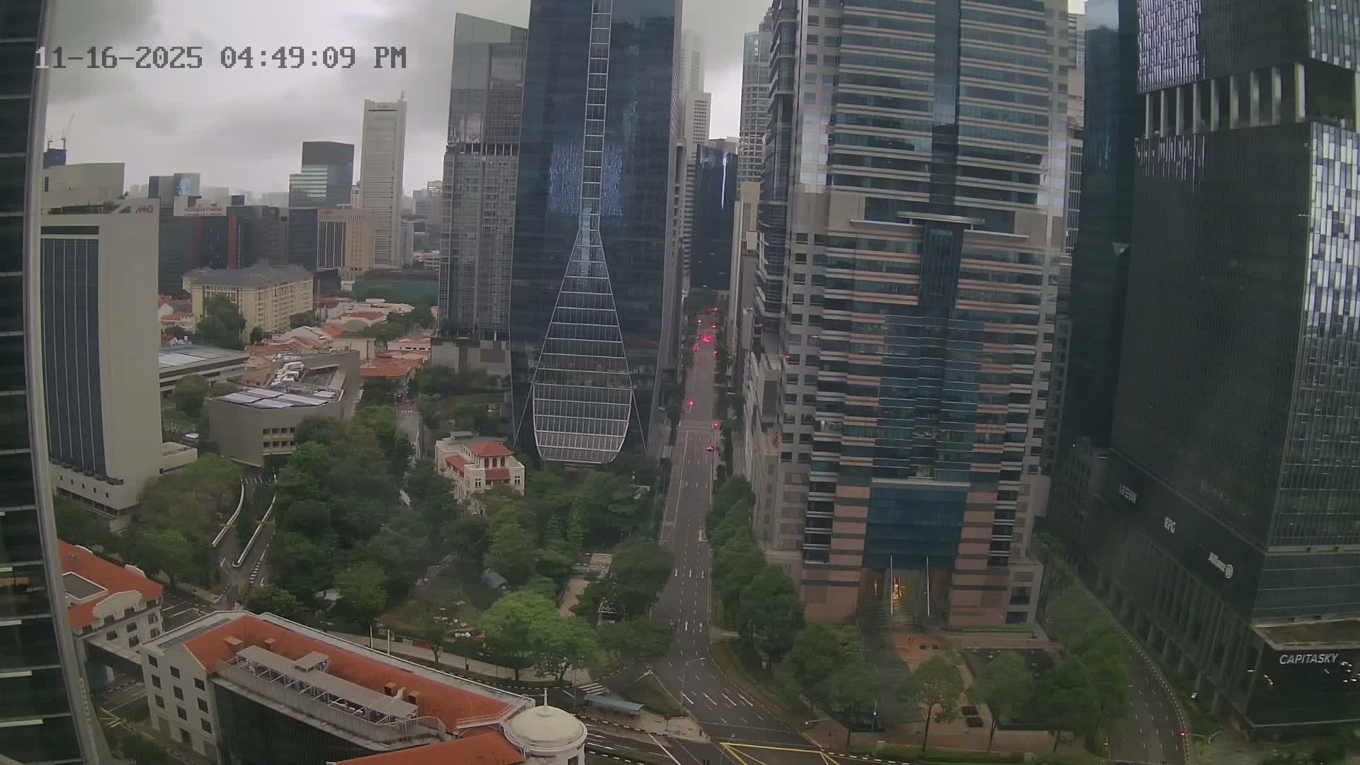 A wide-angle view from above captures a dense urban environment with a mix of towering glass skyscrapers and lower-rise structures, winding roads showing faint traffic lights, and patches of green, all under a heavily overcast and gloomy sky.