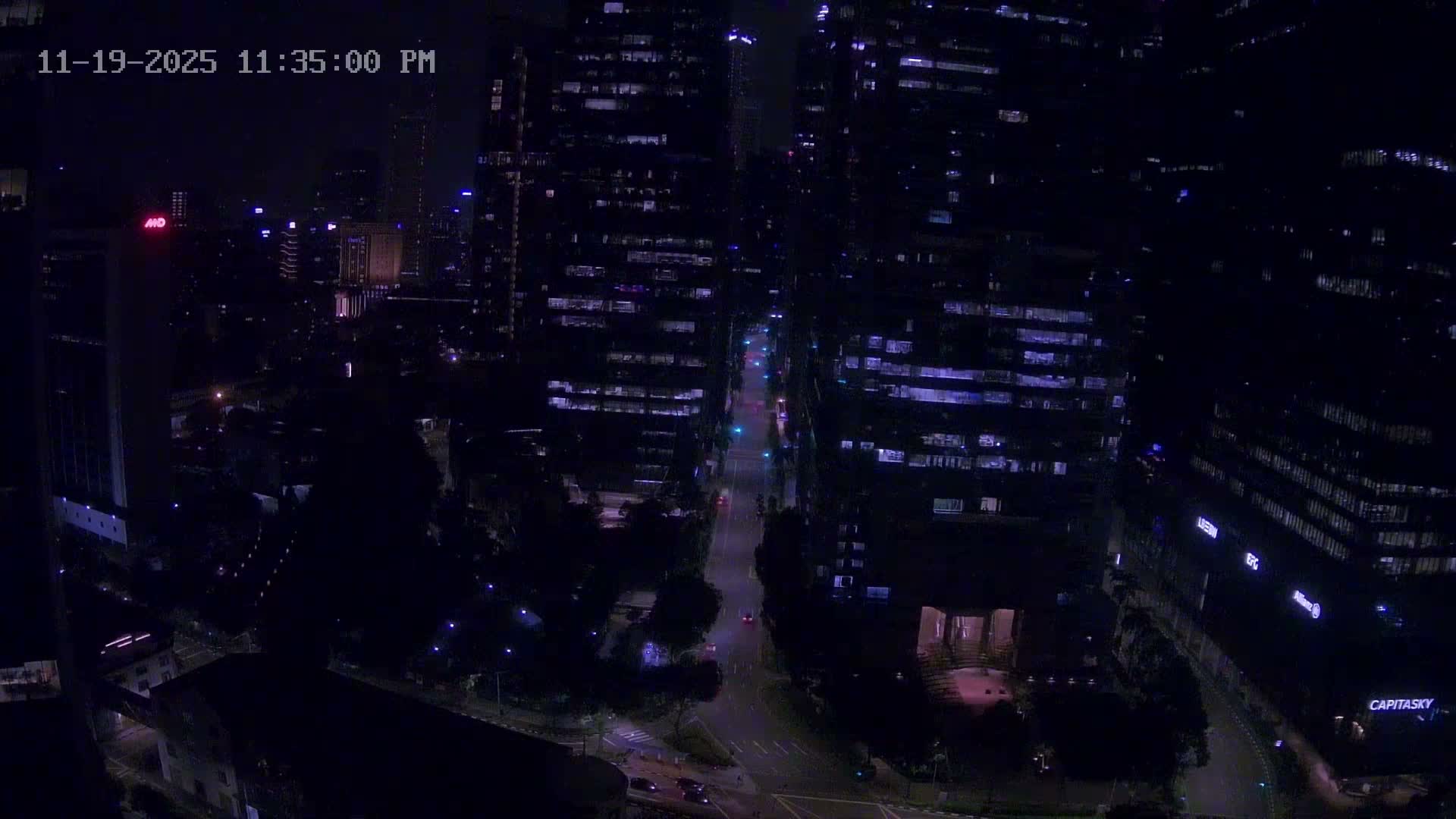 A wide-angle view from above captures a dense urban environment with a mix of towering glass skyscrapers and lower-rise structures, winding roads showing faint traffic lights, and patches of green, all under a heavily overcast and gloomy sky.