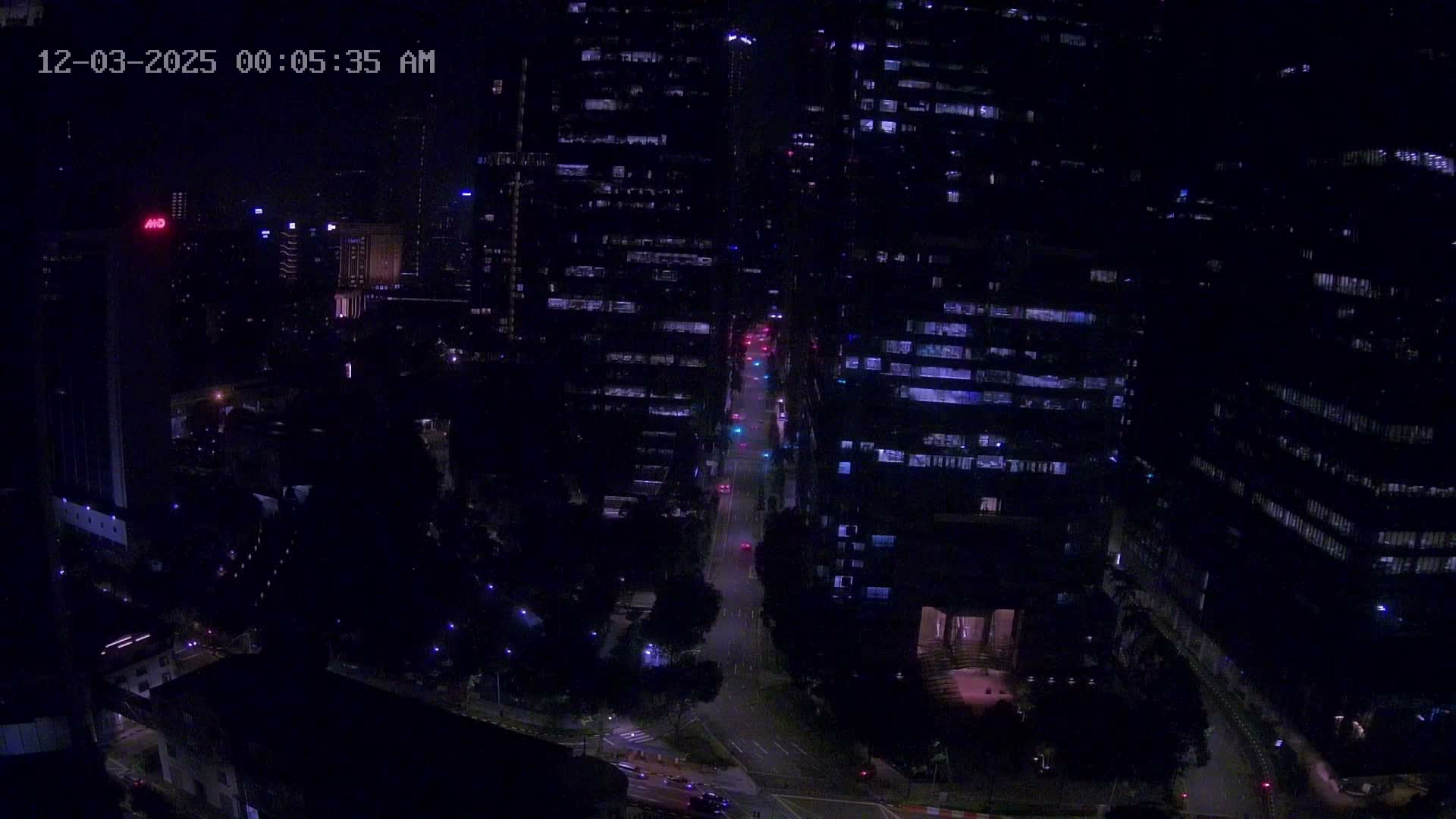 An aerial view captures a vibrant city at night, with multiple high-rise buildings displaying illuminated windows and a central street bustling with vehicles under traffic lights, all beneath a clear, dark sky.