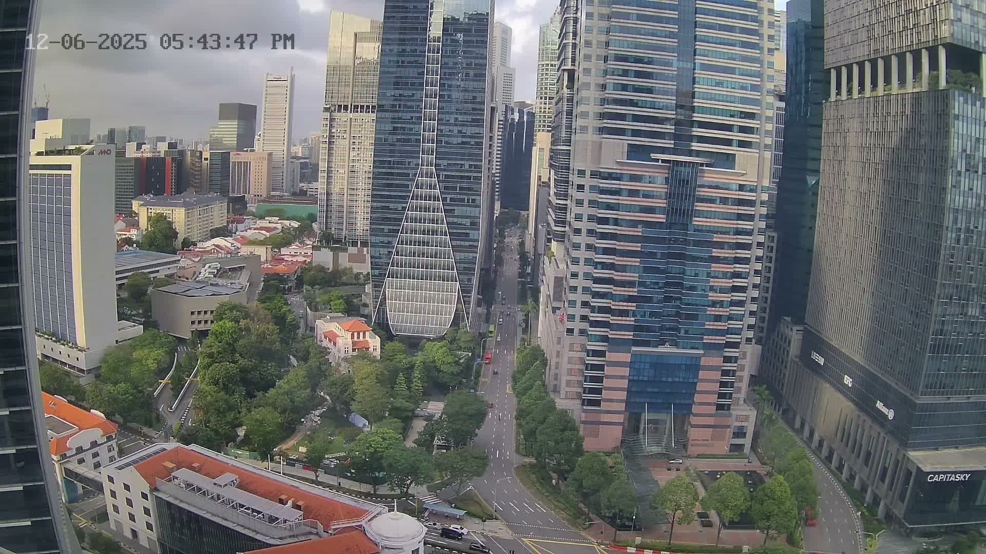 An aerial view captures a bustling urban landscape featuring numerous tall modern skyscrapers alongside lower-rise buildings and abundant green spaces, all under an overcast sky.