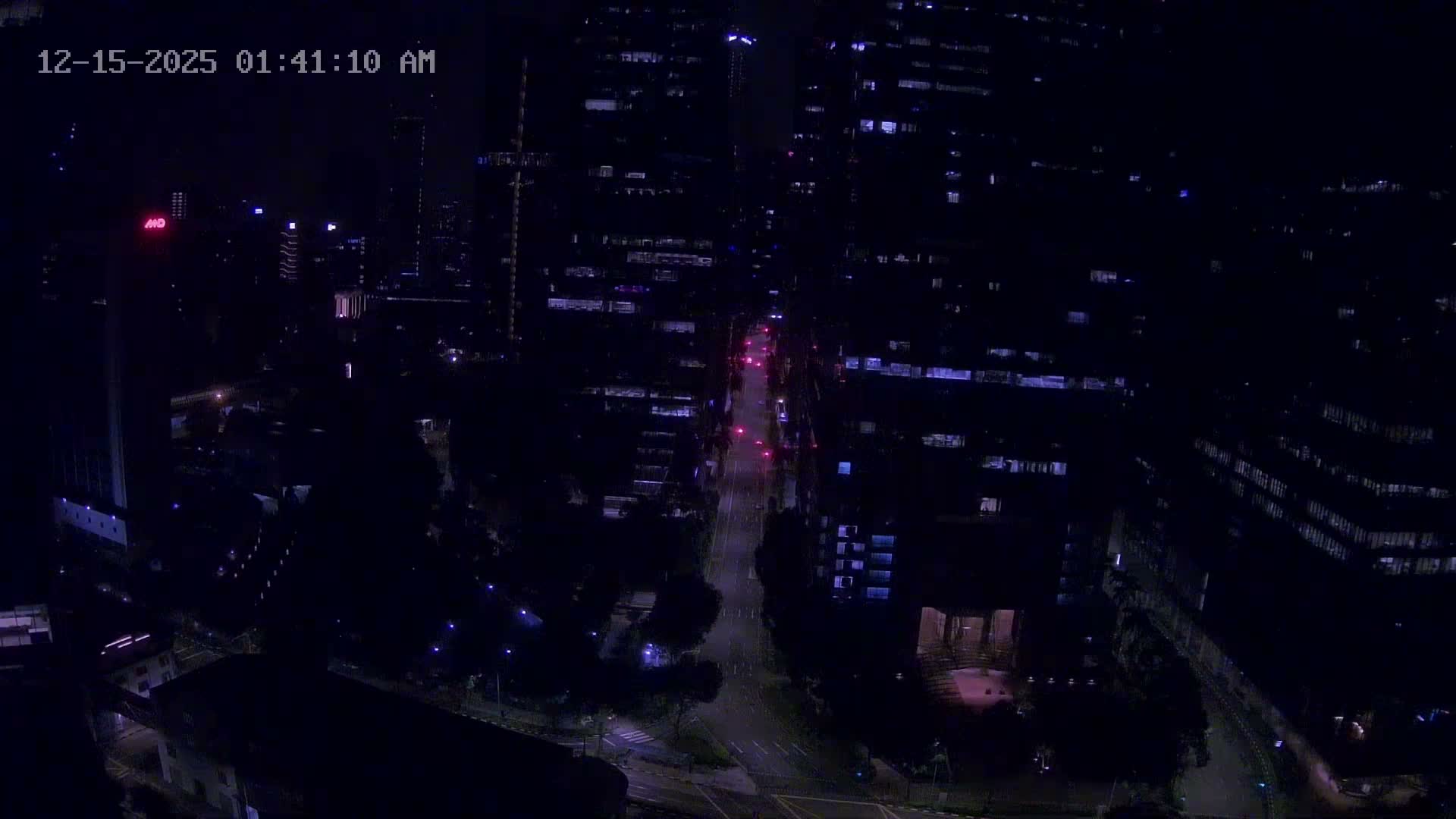 An aerial view captures a bustling urban landscape featuring numerous tall modern skyscrapers alongside lower-rise buildings and abundant green spaces, all under an overcast sky.