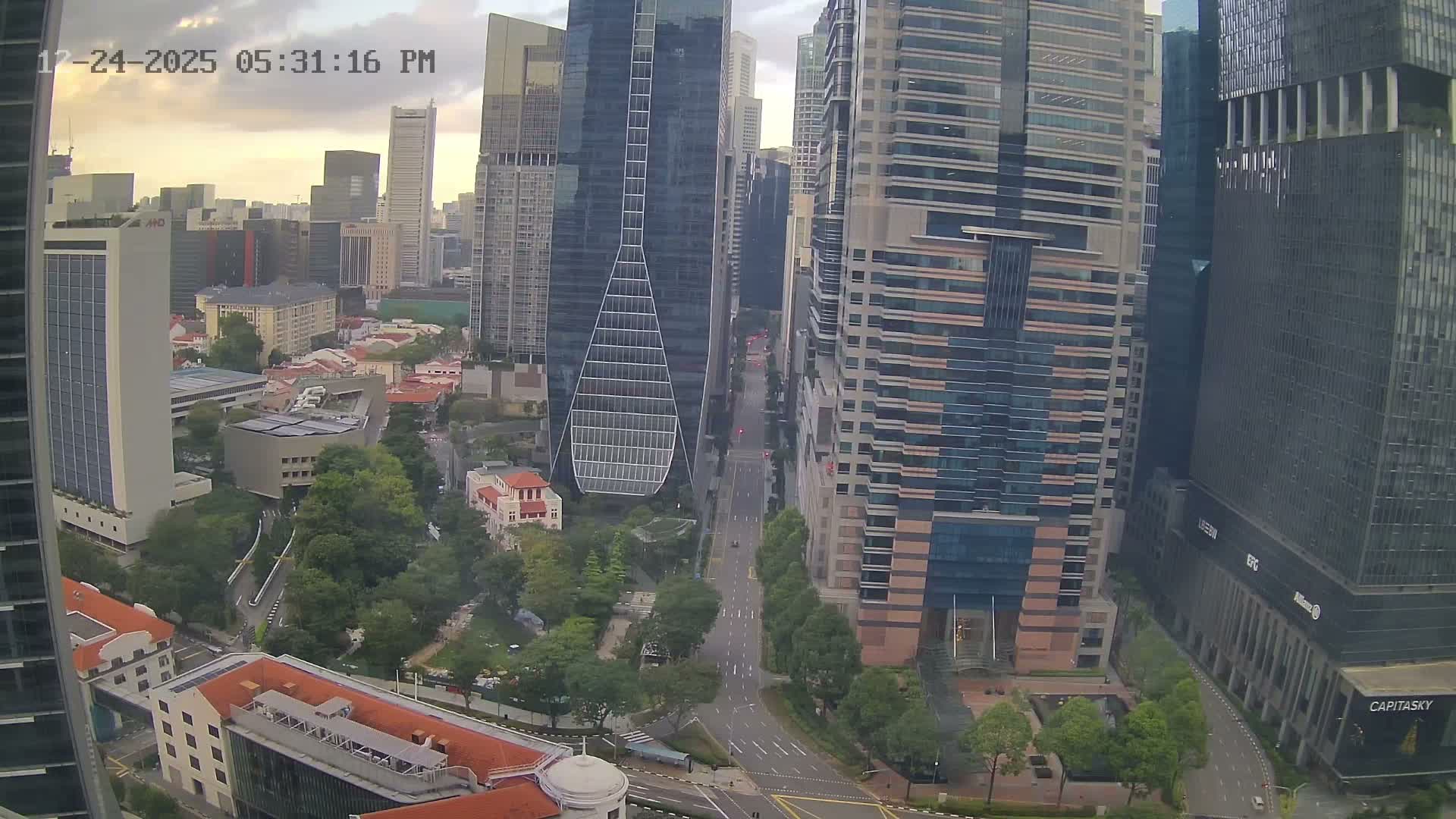 An aerial view captures a bustling urban landscape featuring numerous tall modern skyscrapers alongside lower-rise buildings and abundant green spaces, all under an overcast sky.