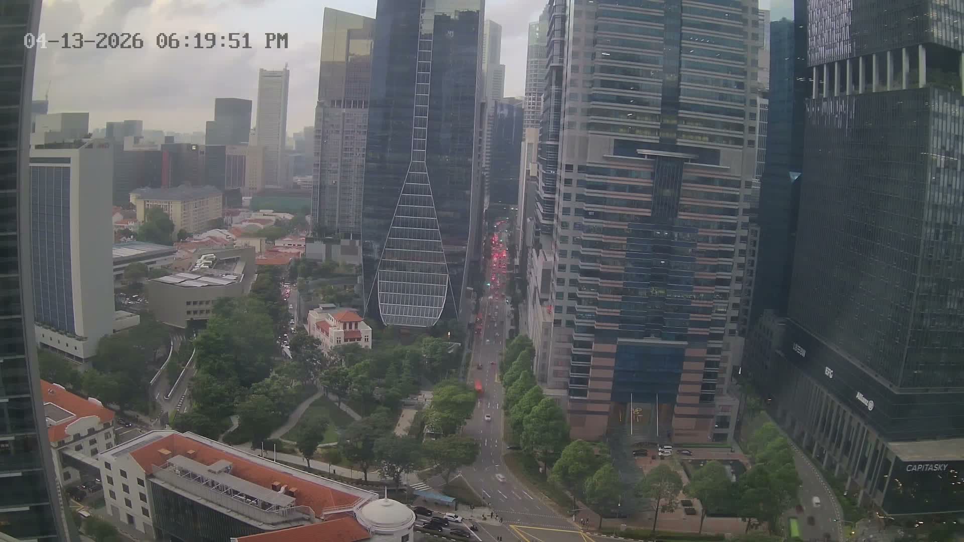 An aerial view captures a bustling urban landscape featuring numerous tall modern skyscrapers alongside lower-rise buildings and abundant green spaces, all under an overcast sky.
