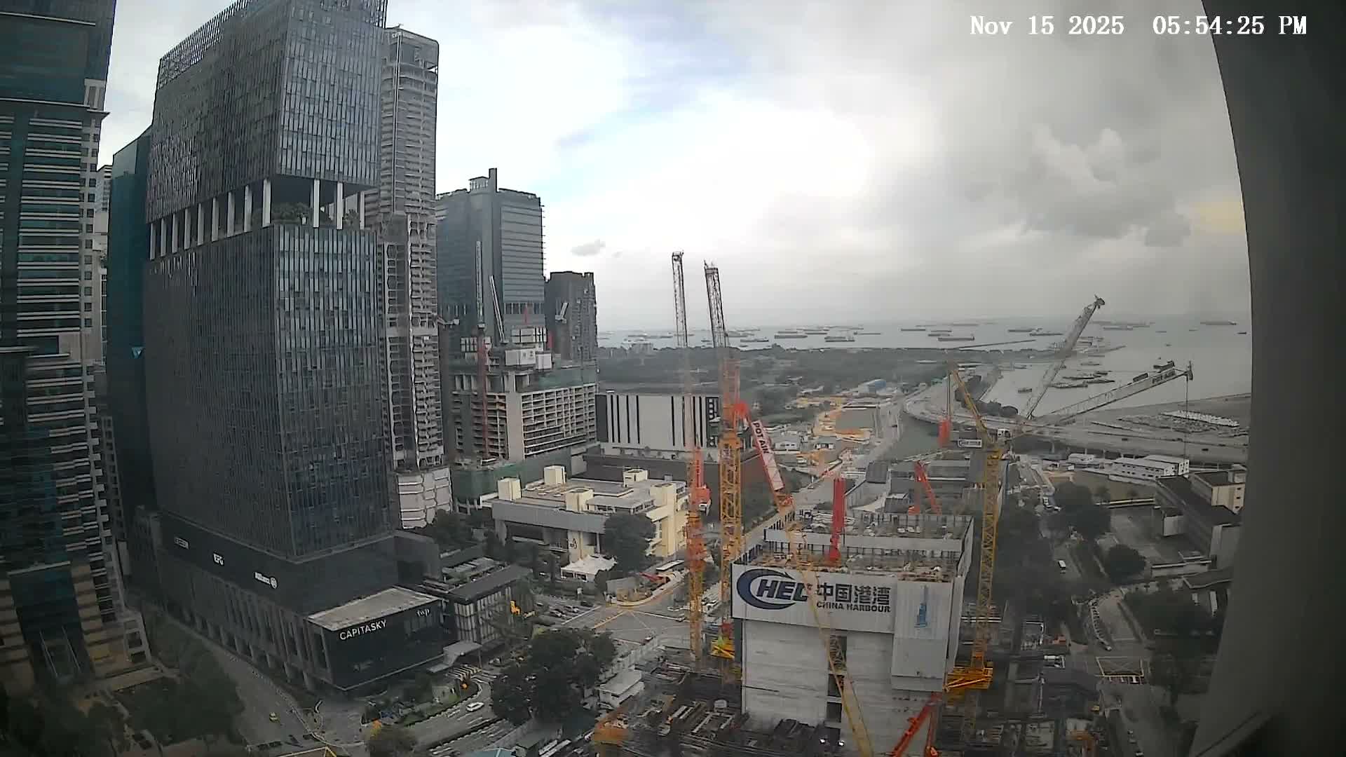 A vast cityscape featuring numerous skyscrapers and ongoing construction with many towering cranes overlooks a busy bay filled with cargo ships under a cloudy, late afternoon sky.