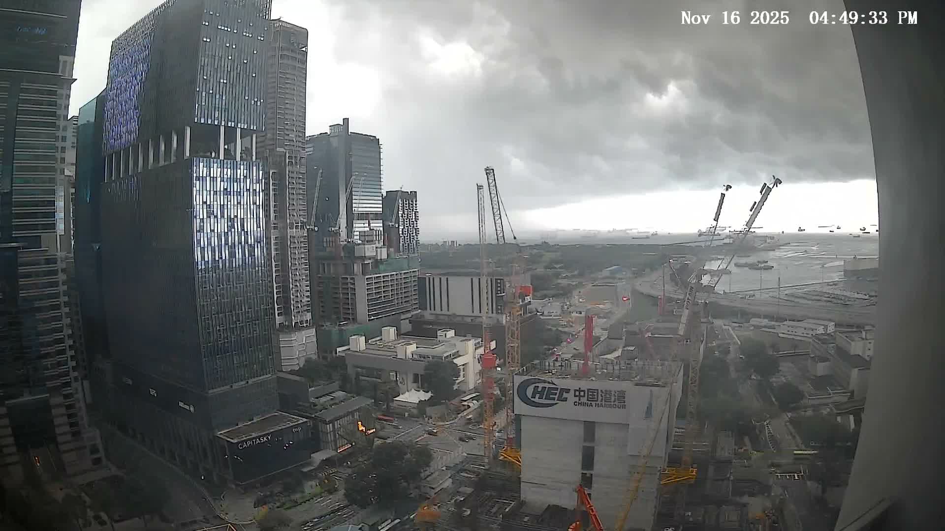 A sprawling urban landscape is depicted with numerous modern skyscrapers, several construction sites featuring tall cranes, and a distant maritime area filled with ships, all beneath a dense, overcast, and gloomy sky.