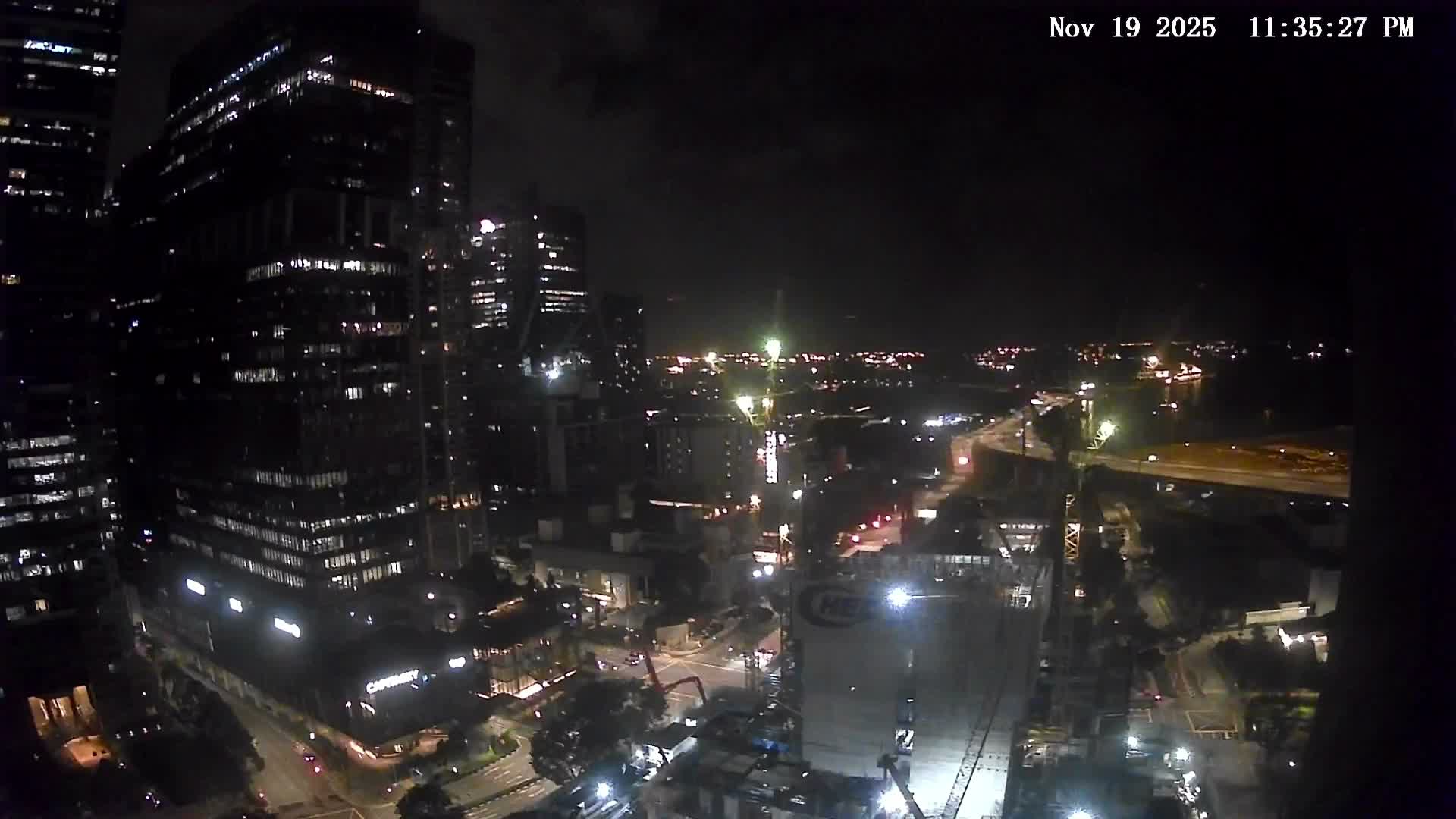 A sprawling urban landscape is depicted with numerous modern skyscrapers, several construction sites featuring tall cranes, and a distant maritime area filled with ships, all beneath a dense, overcast, and gloomy sky.