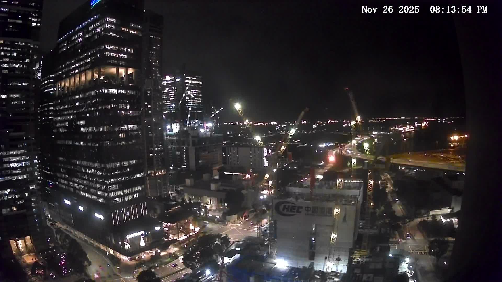 An overcast sky hangs over a dense urban landscape featuring numerous skyscrapers, active construction sites with multiple cranes, and a bustling harbor dotted with many ships.