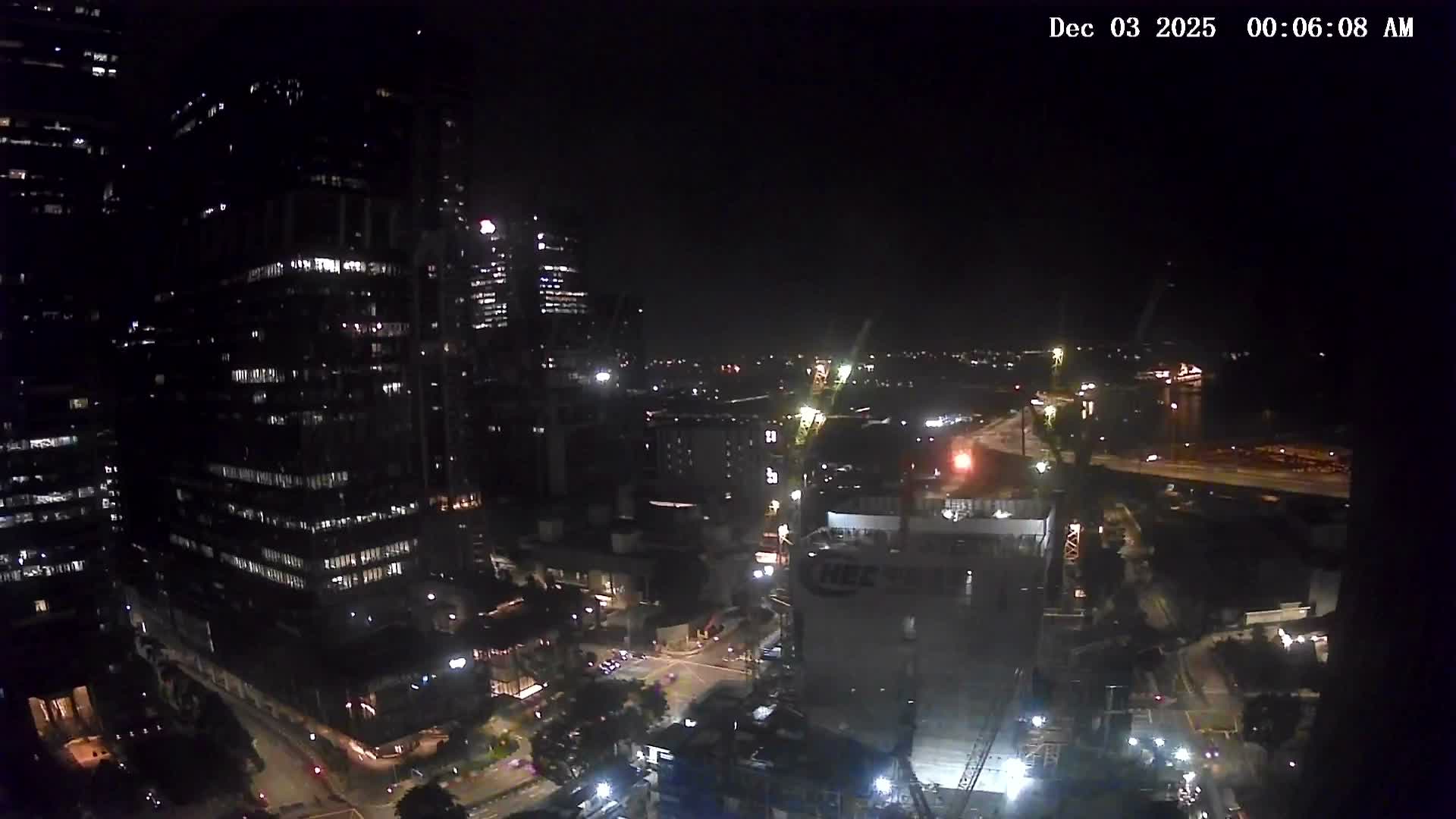 An elevated nighttime view reveals a sprawling cityscape with brightly lit high-rise buildings, busy streets, and multiple construction sites illuminated under a clear, dark sky.