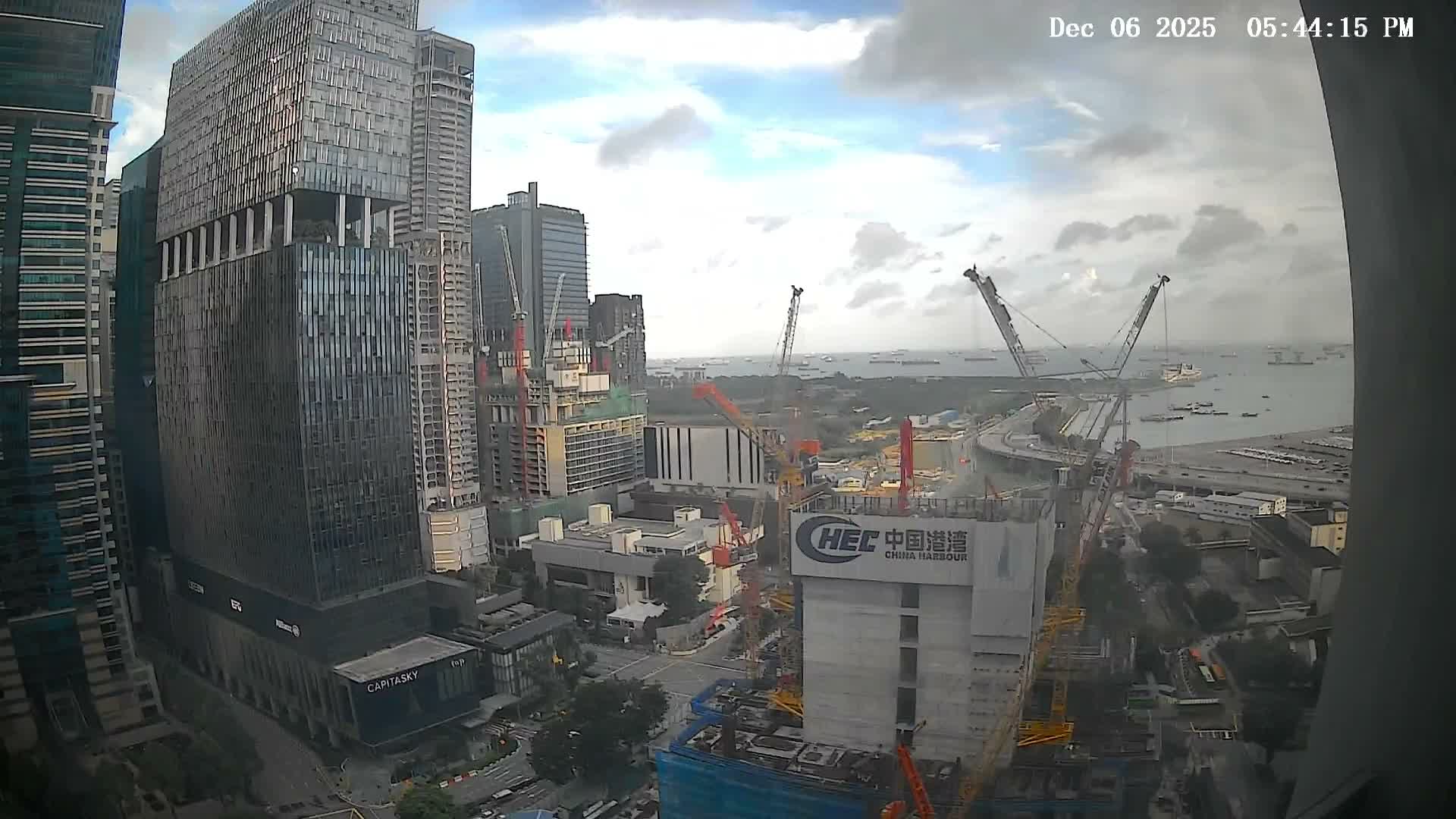 Numerous skyscrapers and active construction sites with multiple cranes dominate an urban landscape that overlooks a busy harbor filled with ships under a partly cloudy sky.