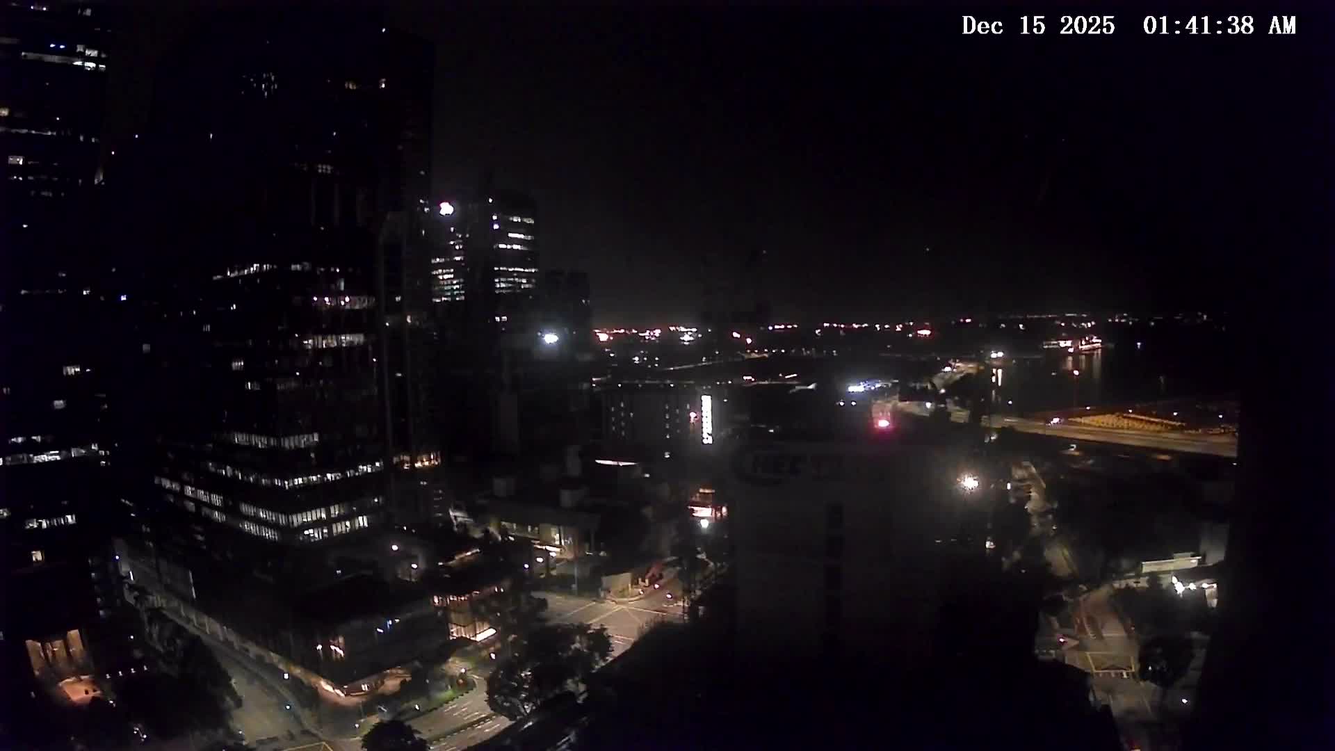 Numerous skyscrapers and active construction sites with multiple cranes dominate an urban landscape that overlooks a busy harbor filled with ships under a partly cloudy sky.