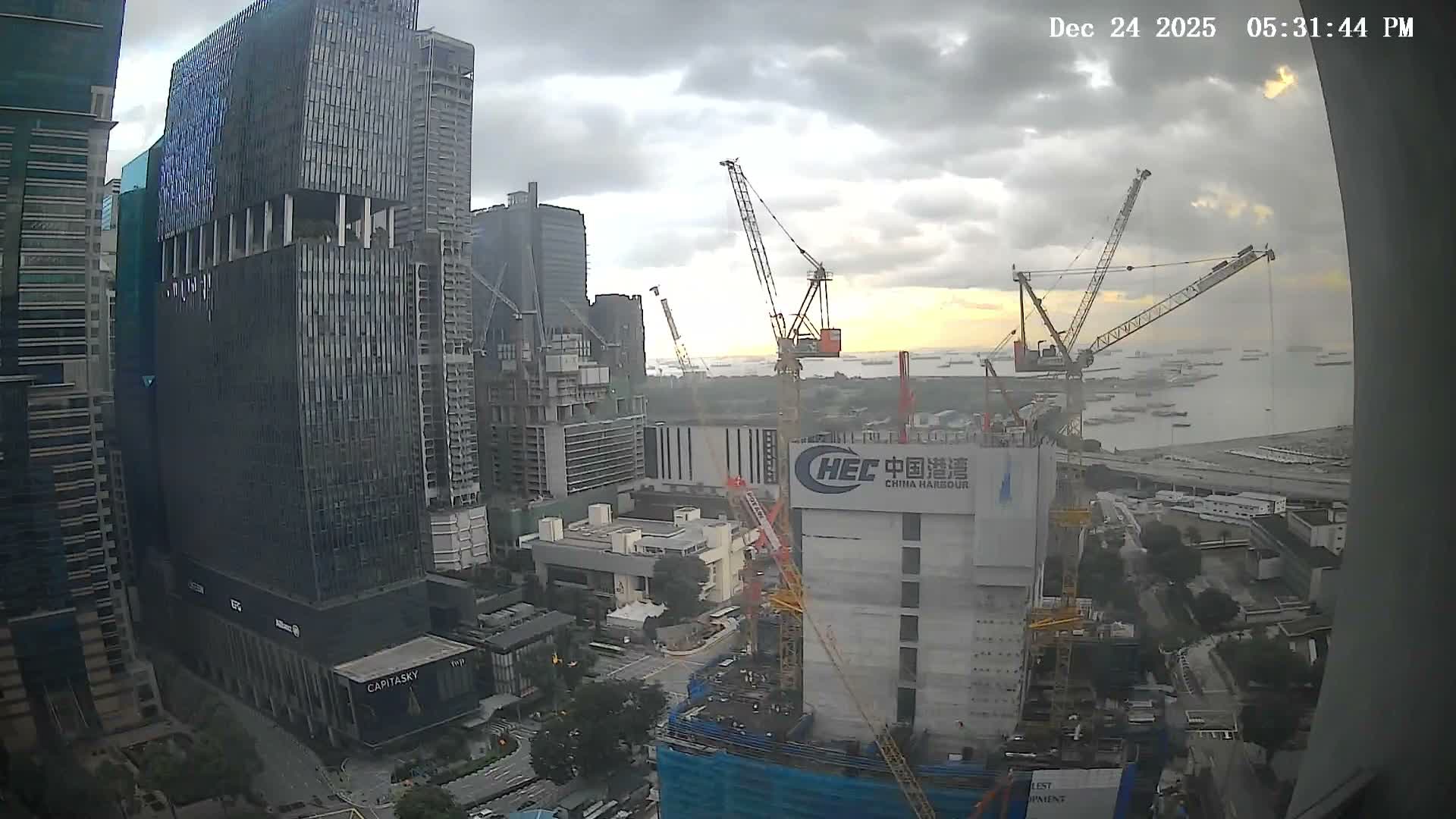 Numerous skyscrapers and active construction sites with multiple cranes dominate an urban landscape that overlooks a busy harbor filled with ships under a partly cloudy sky.